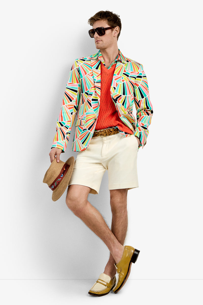A man wearing the THURSTON BLAZER with a multicolored geometric pattern, sunglasses, an orange sweater, cream shorts, a brown belt, and yellow loafers holds a straw hat against a plain white background.