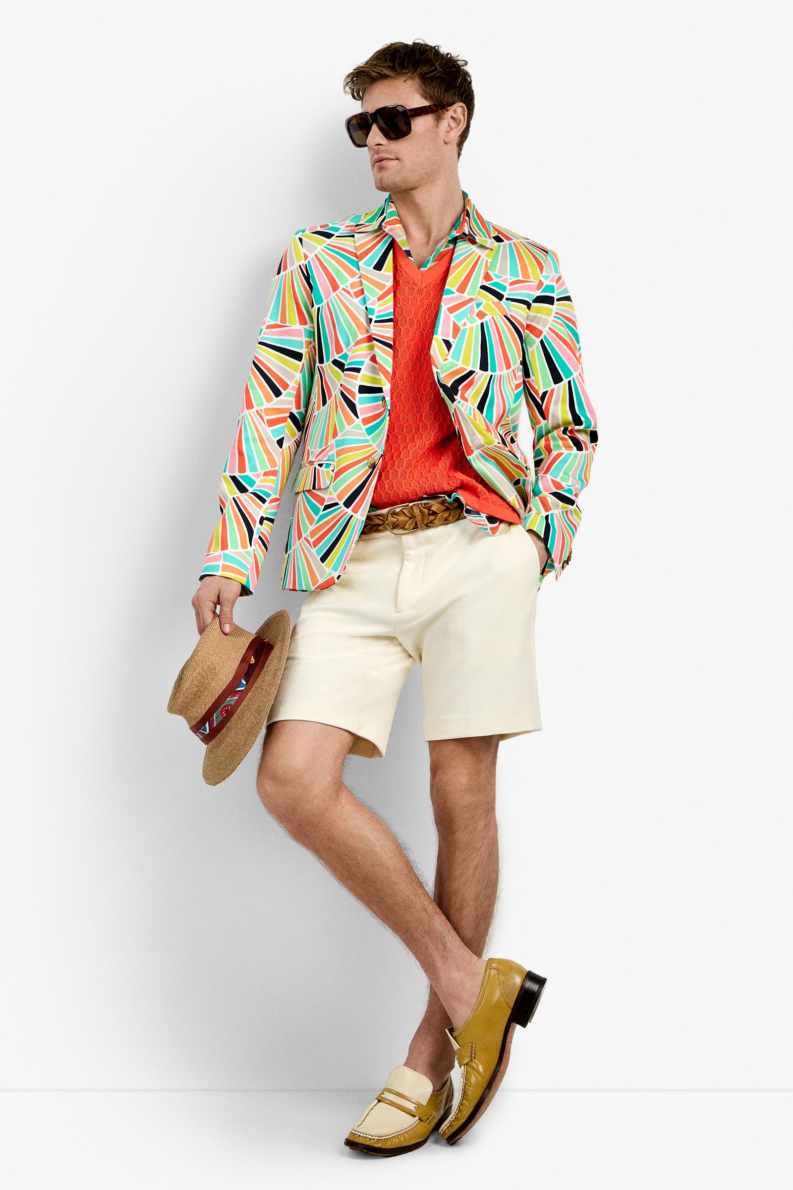 A man wearing the THURSTON BLAZER with a multicolored geometric pattern, sunglasses, an orange sweater, cream shorts, a brown belt, and yellow loafers holds a straw hat against a plain white background.