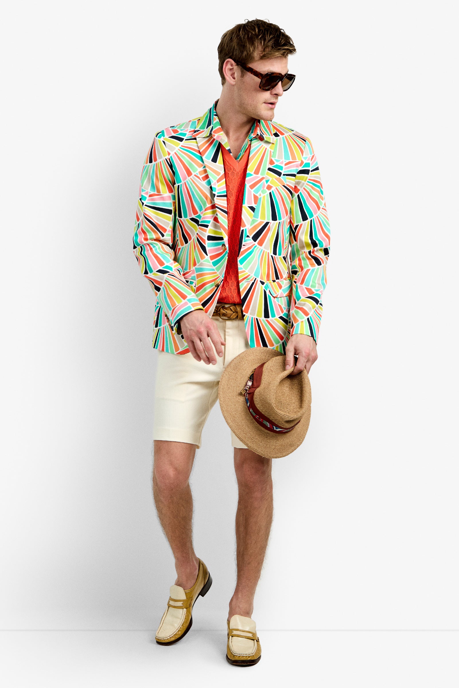 A man in sunglasses, a THURSTON BLAZER with colorful geometric patterns, a red shirt, white shorts, brown belt, and beige loafers stands against a white background, holding a tan hat with a brown band by his side.