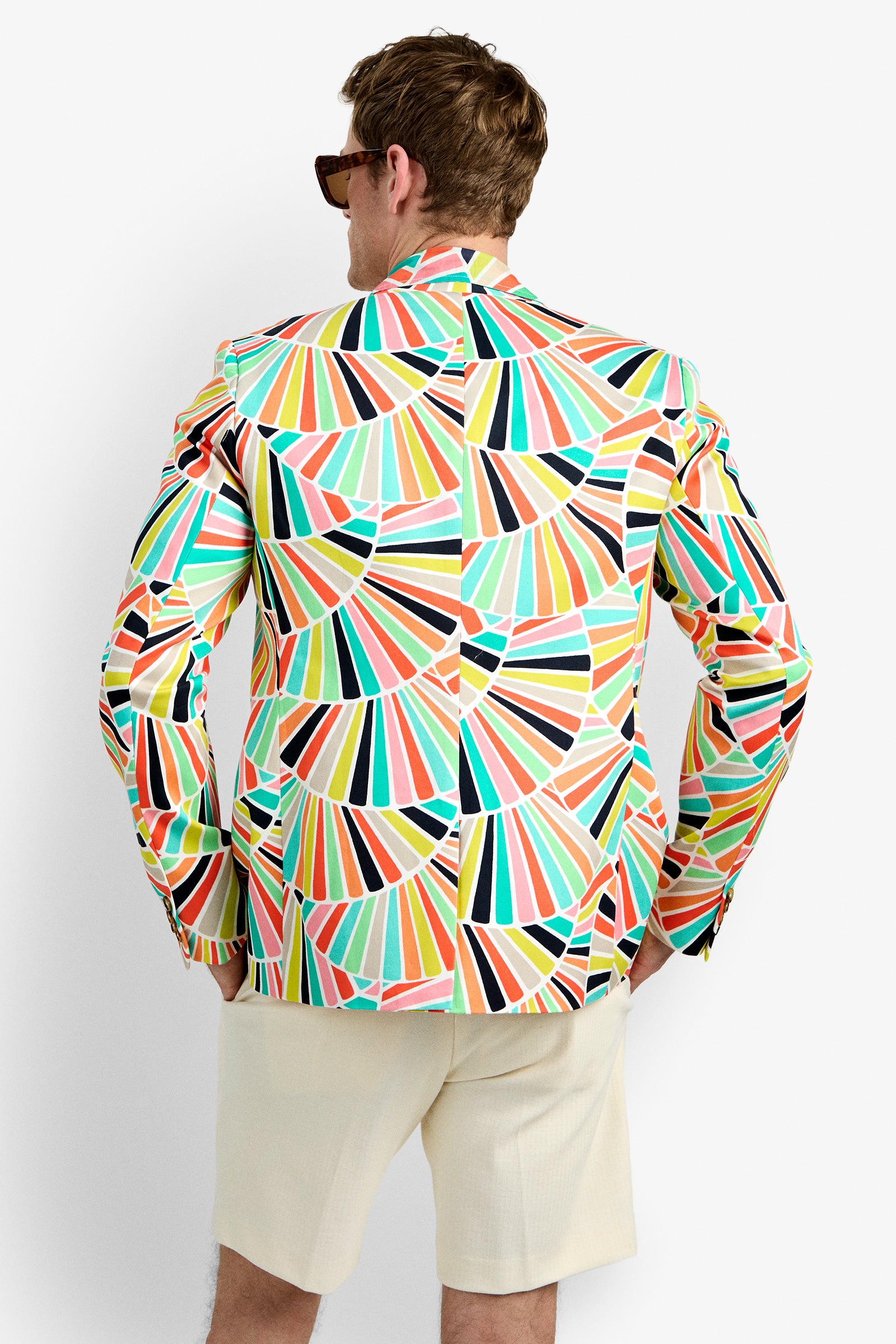 A man faces away from the camera, wearing sunglasses, cream-colored shorts, and the THURSTON BLAZER featuring a colorful fan pattern, set against a plain white background.
