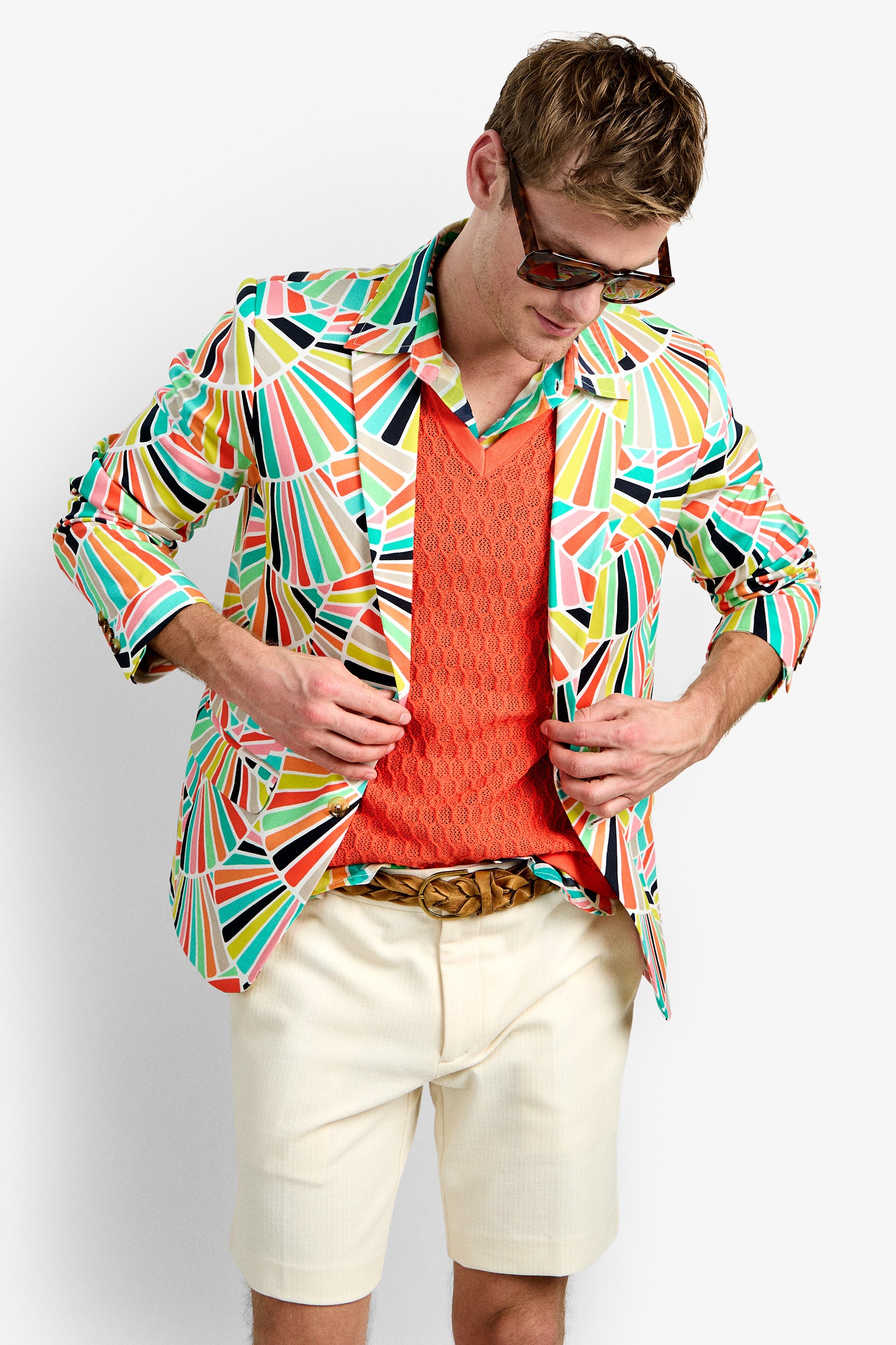 A man in the THURSTON BLAZER with a colorful, geometric pattern, paired with an orange textured sweater, beige shorts, a braided belt, and sunglasses looks down while adjusting his jacket.