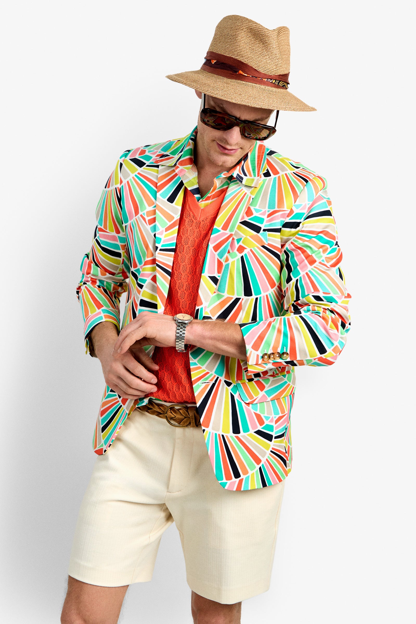 A man wearing the THURSTON BLAZER with a colorful geometric pattern, orange shirt, beige shorts, straw hat with red band, sunglasses, and a watch poses with his head tilted down and hands adjusting his belt.