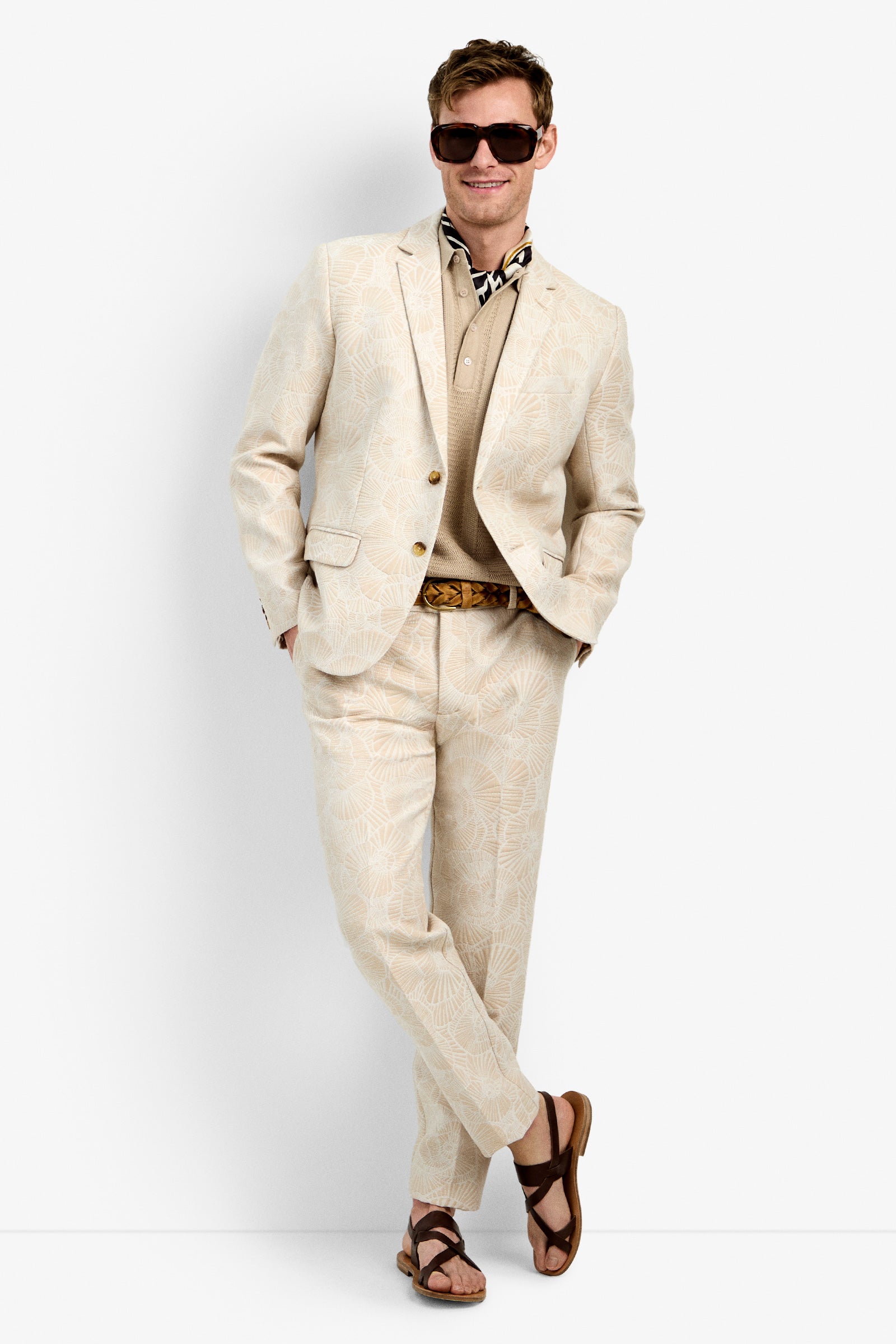 A man stands against a white background wearing the Thurtson Blazer, a light beige patterned suit, along with a tan shirt, brown belt, patterned scarf, sunglasses, and brown sandals. He smiles with one leg crossed in front of the other.