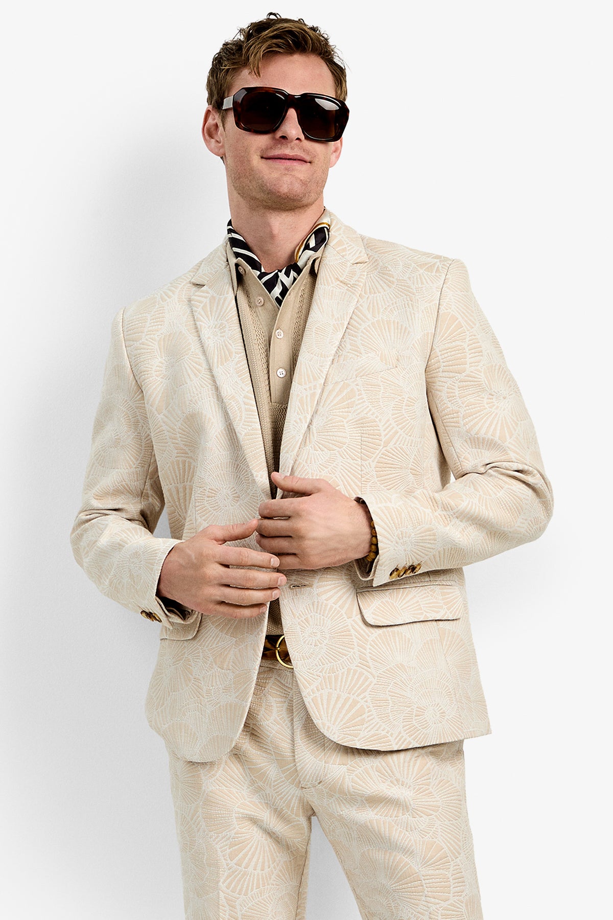 A man wears the Thurtson Blazer—a cream-colored patterned suit—paired with a beige shirt, patterned neck scarf, and dark sunglasses, standing confidently against a plain white background.