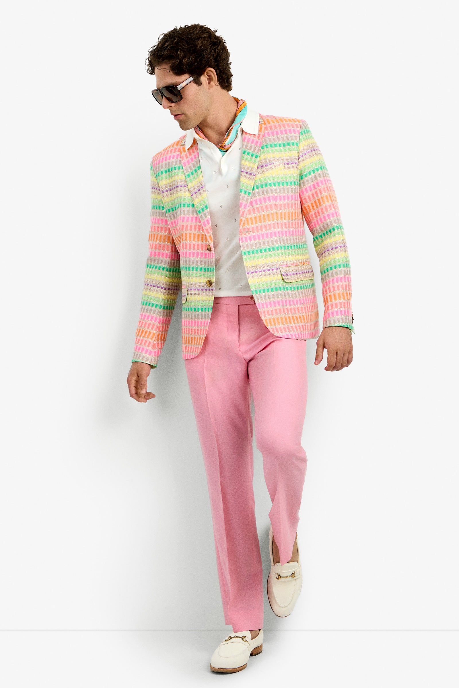 A man in sunglasses, a white shirt, the THURSTON BLAZER in MULTI, light pink pants, and white loafers stands against a plain white background, looking down and to the side.