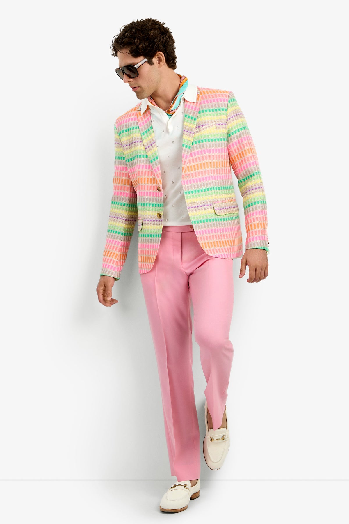 A man in sunglasses, a white shirt, pink pants, white loafers, and the Thurston Blazer—featuring a pastel plaid pattern in pink, yellow, blue, and green—stands against a plain white background.