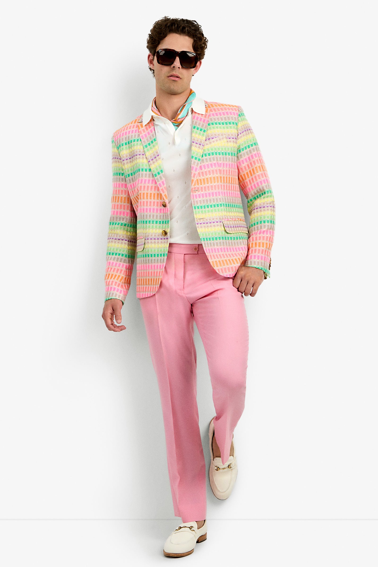 A man in sunglasses, a THURSTON BLAZER with pastel pink, green, orange, and yellow stripes, a white shirt, pink trousers, and cream loafers stands against a white background.