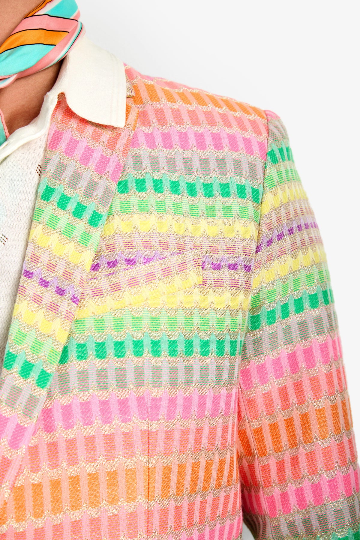 A close-up of a person wearing the Thurston Blazer, featuring colorful stripes of pink, orange, yellow, green, and lavender. They pair it with a white collared shirt and a multicolored striped scarf around their neck.