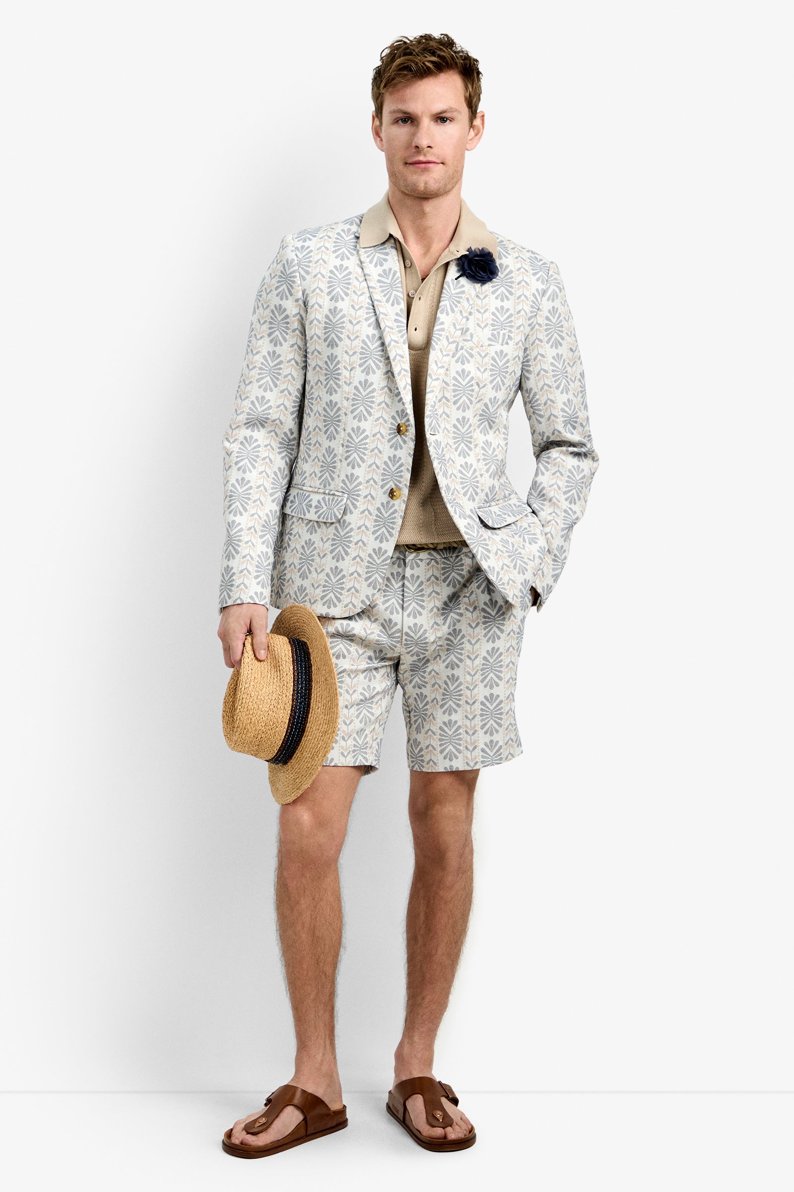 A man wears the Thurston Blazer with matching shorts, a beige shirt, and a dark flower pin. He holds a straw hat and wears brown sandals, standing against a plain white background.