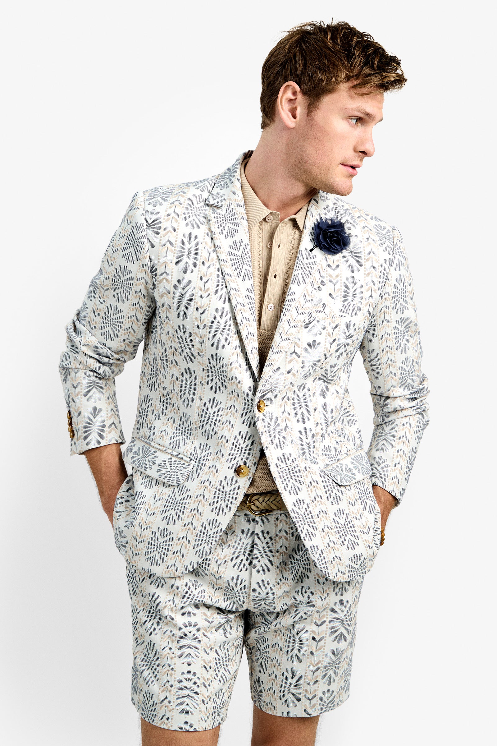 A man stands with hands in pockets, looking to the side against a plain background, wearing the Thurston Blazer and matching shorts over a beige shirt, accessorized with a dark flower pin on the lapel.