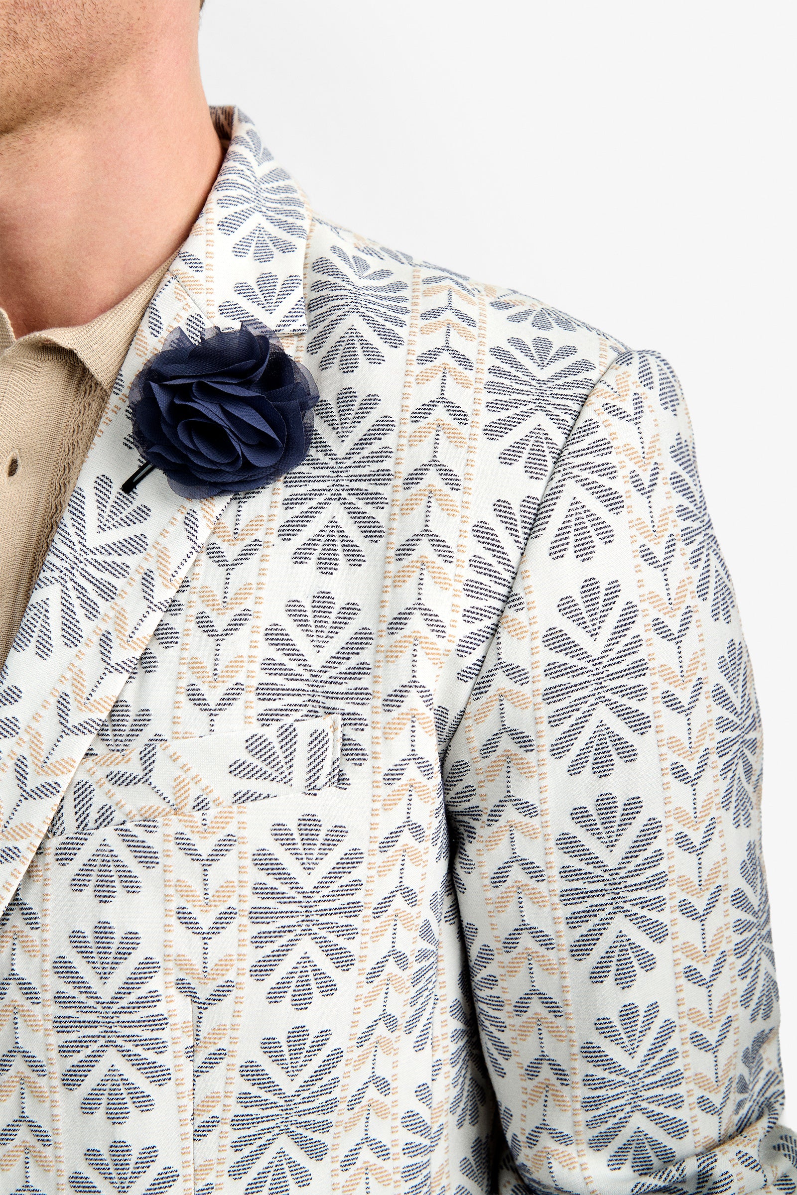 A close-up of a person wearing the Thurston Blazer, featuring blue and beige geometric patterns, over a light beige shirt with a dark blue flower lapel pin. Only part of the person's face is visible.