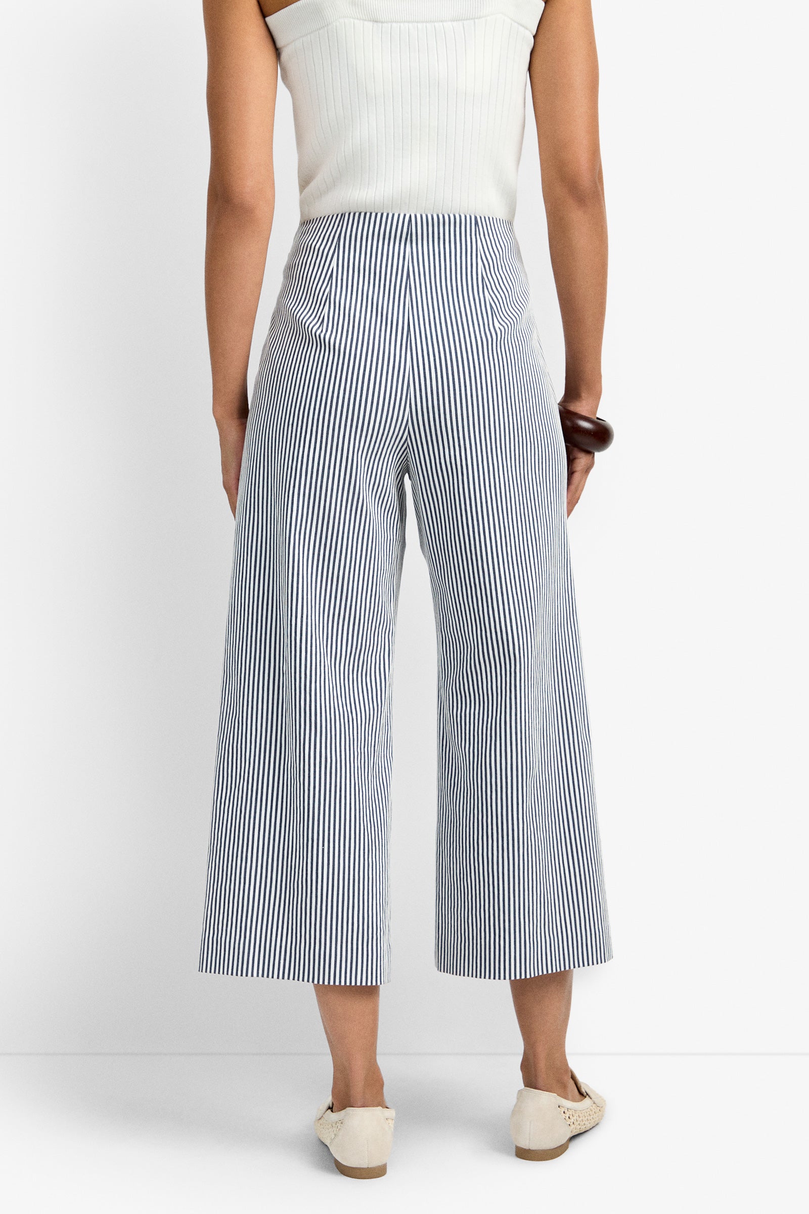 A person is seen from the back wearing a sleeveless white top and the Saltie Pant—high-waisted, wide-leg blue-and-white vertical striped pants with two back pockets—paired with white shoes.