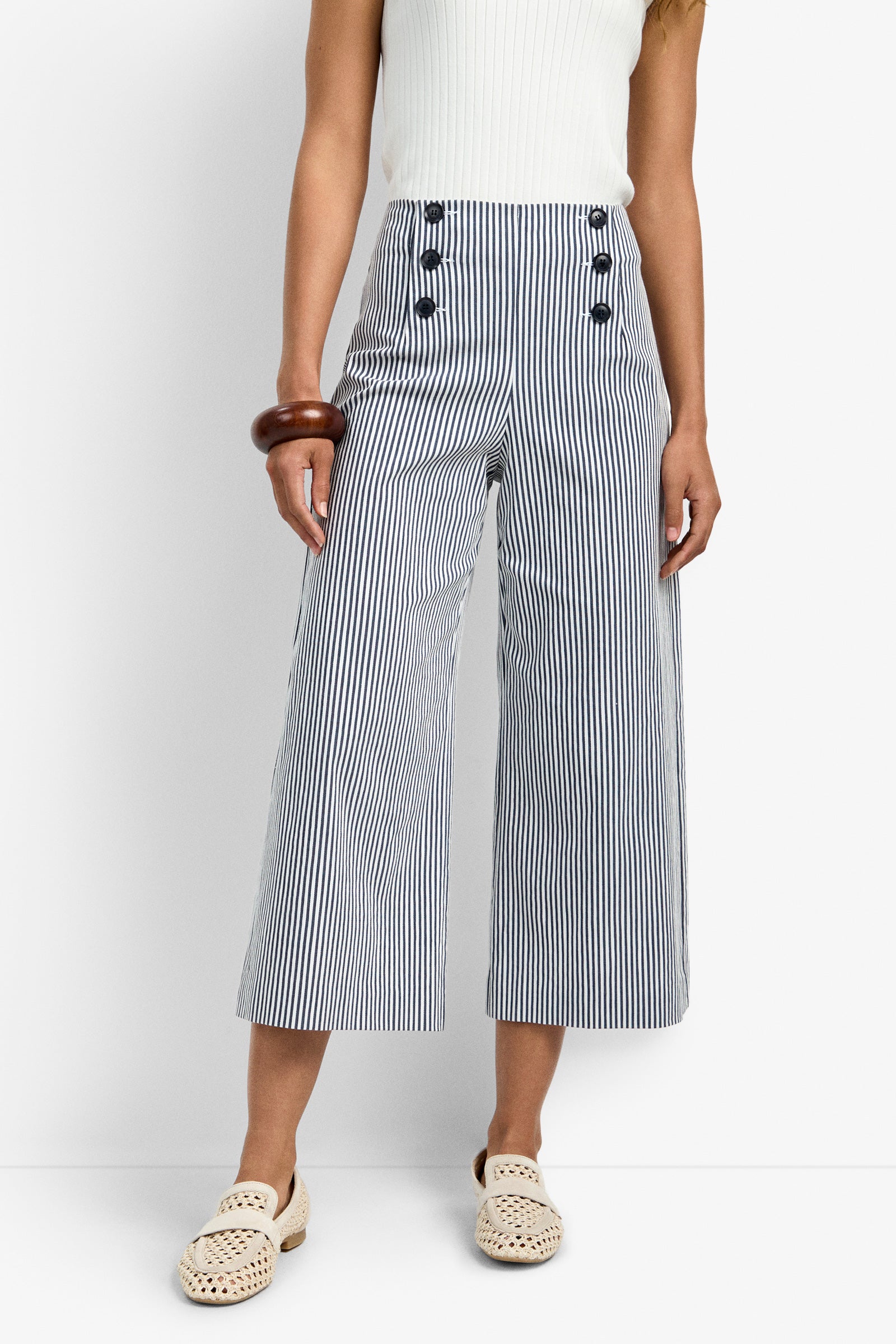 A person models the Saltie Pant—high-waisted, wide-leg, blue-and-white striped pants with large buttons—paired with a sleeveless white top, cream woven shoes, and wooden bangles against a plain background.