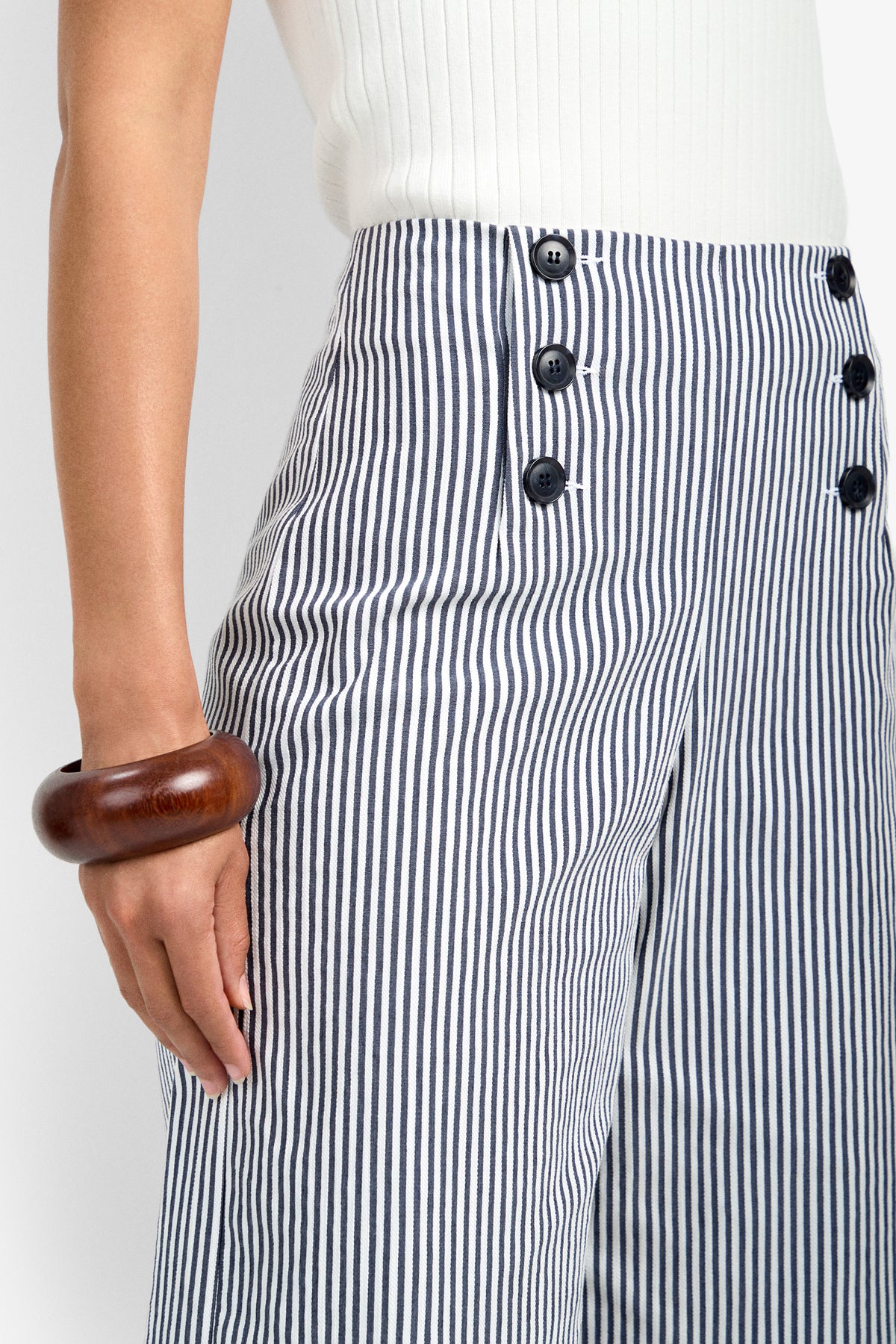 A person wearing the Saltie Pant, featuring high-waisted blue and white vertical stripes with black buttons, paired with a white ribbed top and a chunky wooden bracelet on their left wrist.