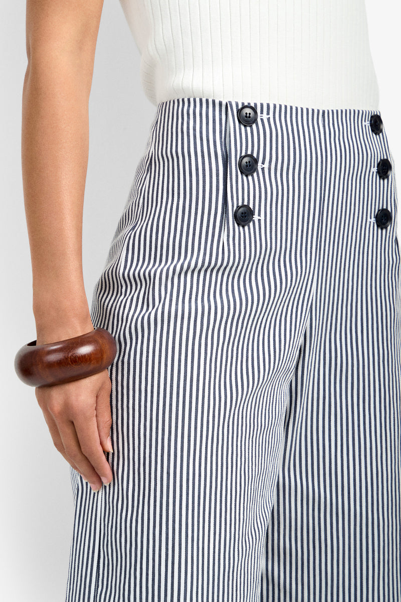 A person wearing the Saltie Pant, featuring high-waisted blue and white vertical stripes with black buttons, paired with a white ribbed top and a chunky wooden bracelet on their left wrist.
