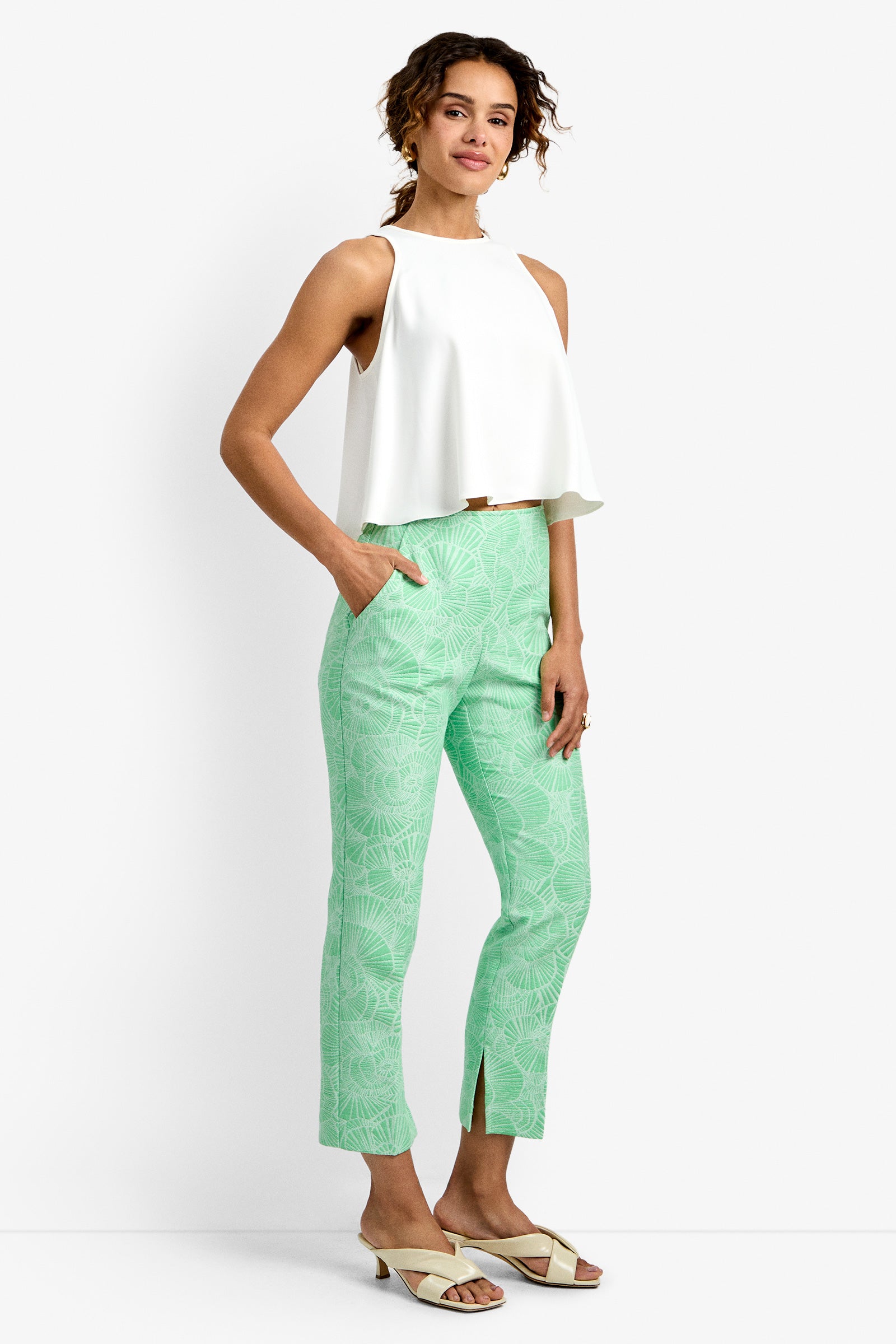 A woman smiles with her hands in her pockets, wearing the Pop Pant—a mint green patterned pant with front slits—paired with a sleeveless white top and white sandals. She has curly hair.