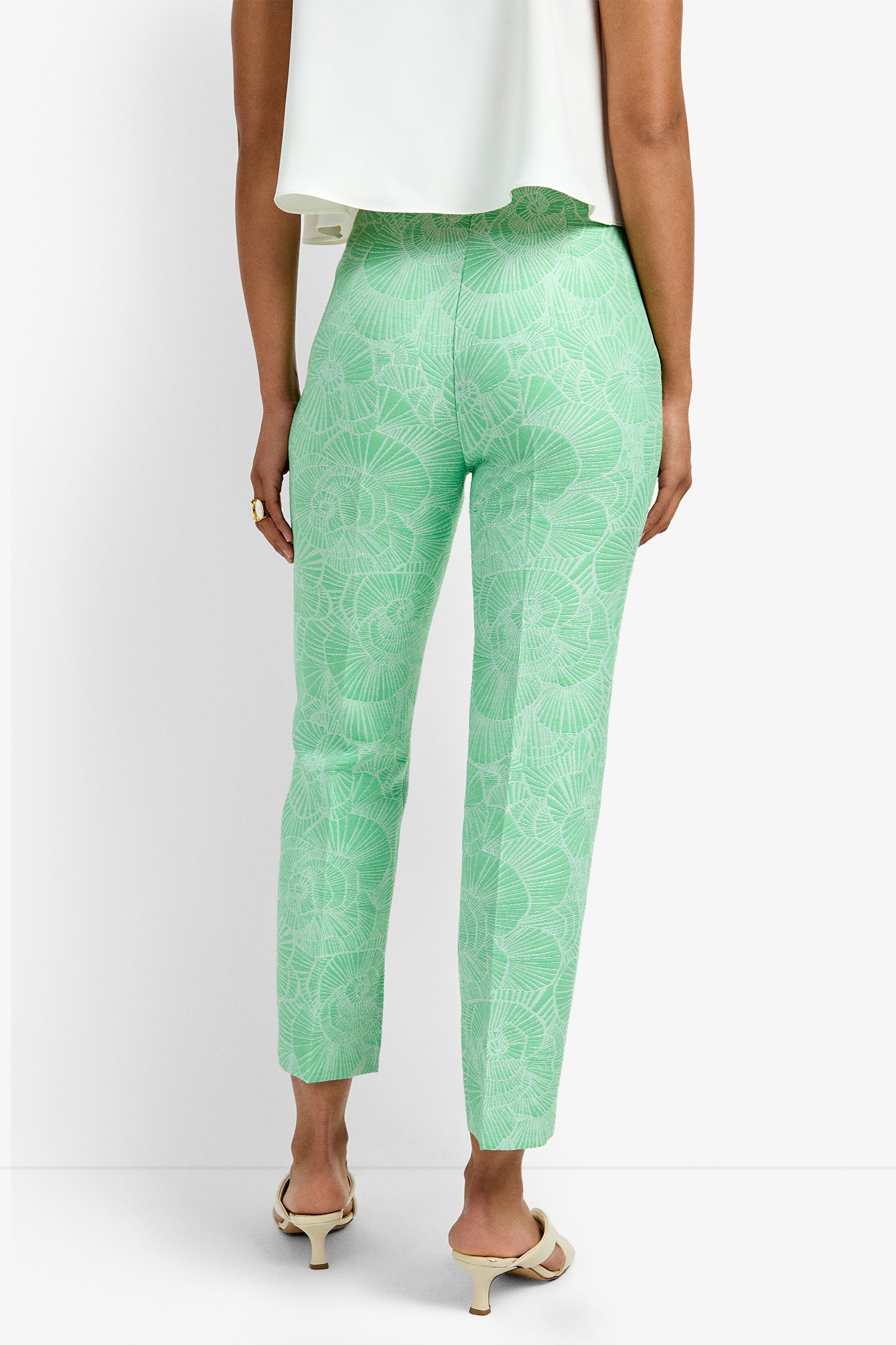 A person stands with their back to the camera wearing Pop Pant in light green with a subtle leaf design, paired with a white top and light heeled sandals.