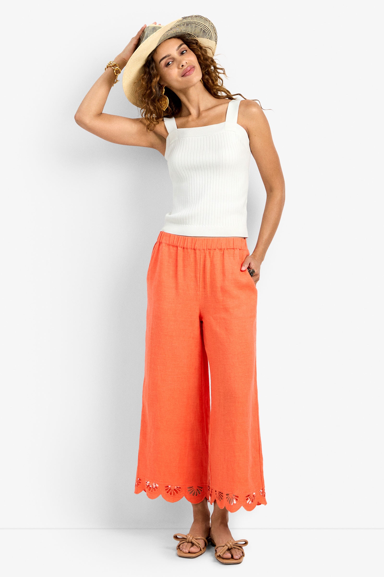 A woman smiles, standing with one hand on her straw hat and the other in her pocket, wearing a white sleeveless ribbed top, coral Cruze Pant with scalloped hems, and brown sandals.