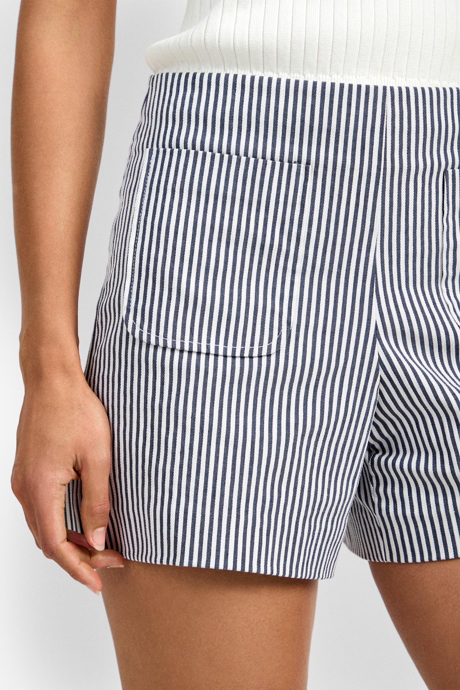 A person wearing the Caspian Short, featuring blue and white vertical stripes, a white ribbed waistband, and a patch pocket, shown from waist to mid-thigh.