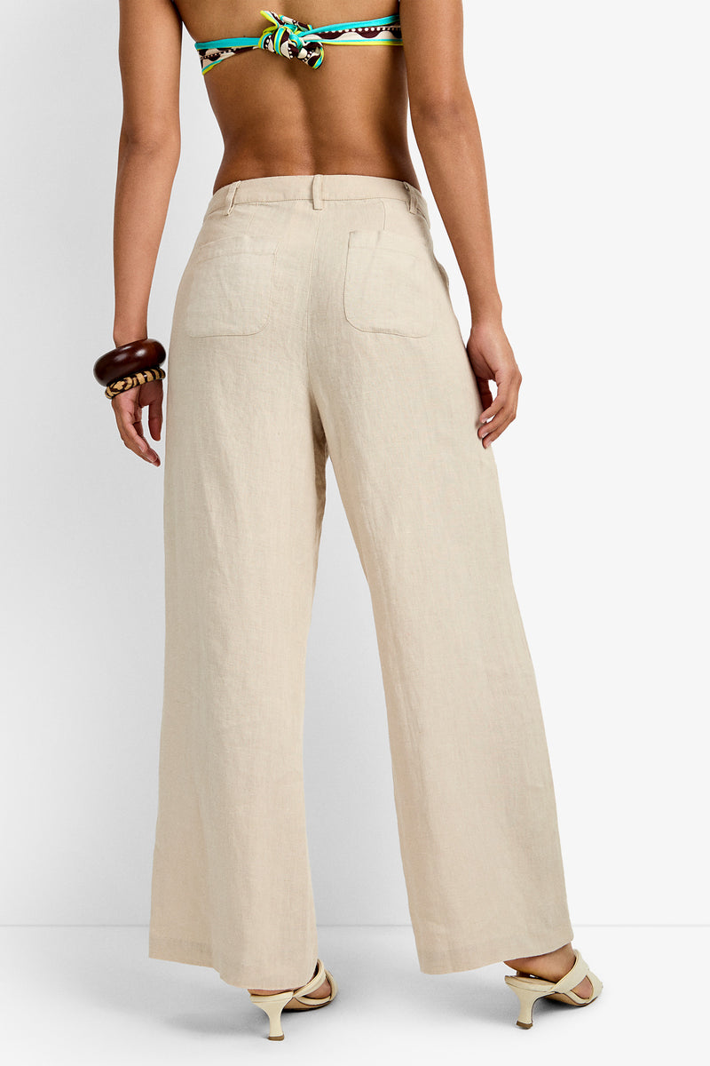 A person is shown from the back wearing the Tonga Pant in beige with a colorful bikini top, chunky brown bracelet, and white heeled sandals.