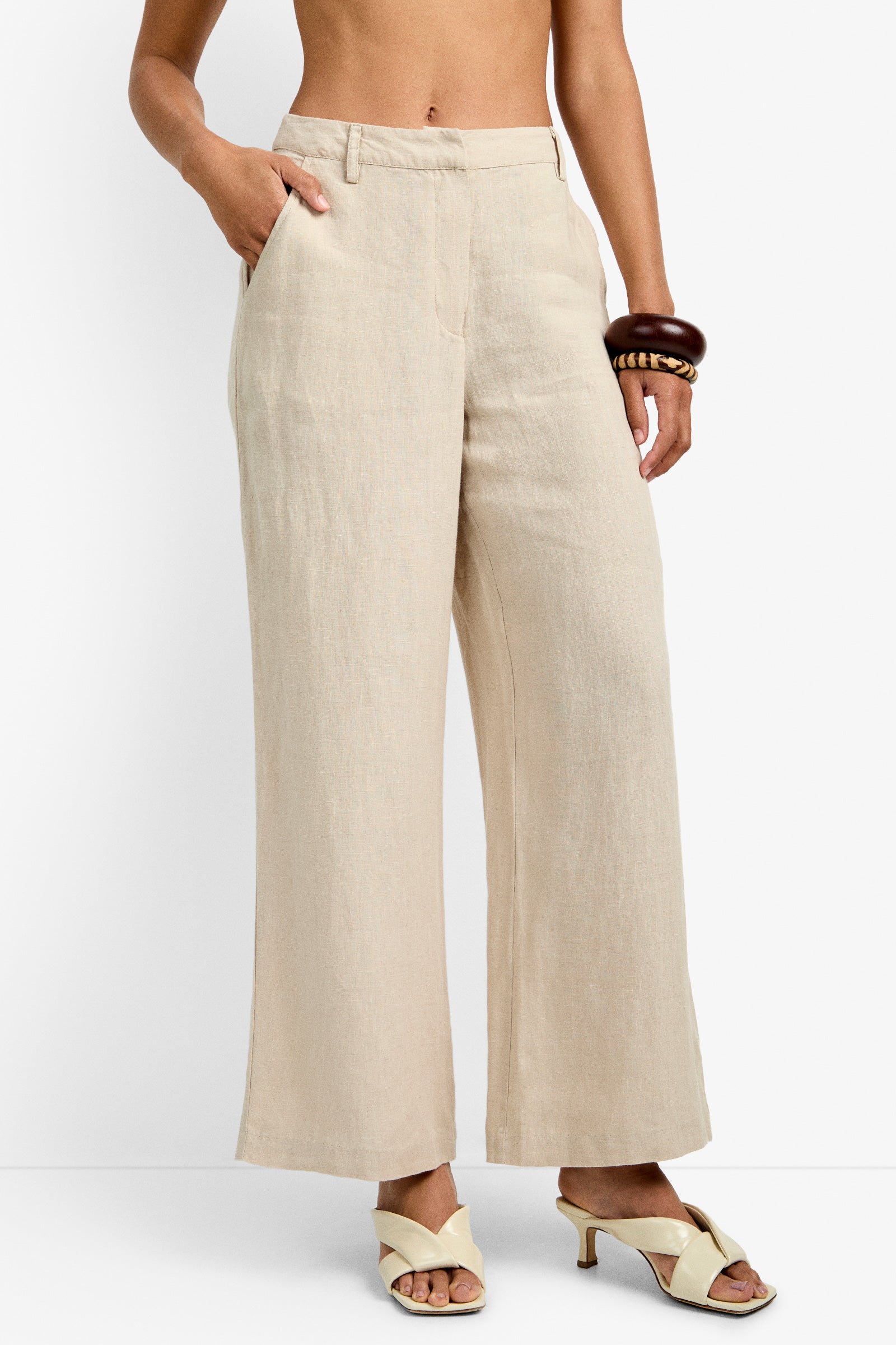 A person wears the Tonga Pant—high-waisted, wide-leg beige linen—with ivory heeled sandals and chunky brown bracelets, one hand in a pocket. Background: plain white. Upper torso is not shown.