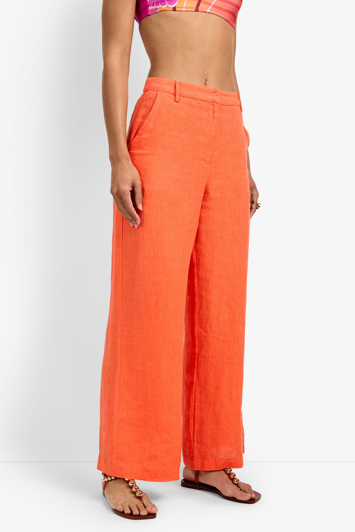 A person wearing high-waisted, wide-leg orange Tonga Pants with a colorful crop top and flat sandals stands against a plain white background. The image is cropped above the shoulders.