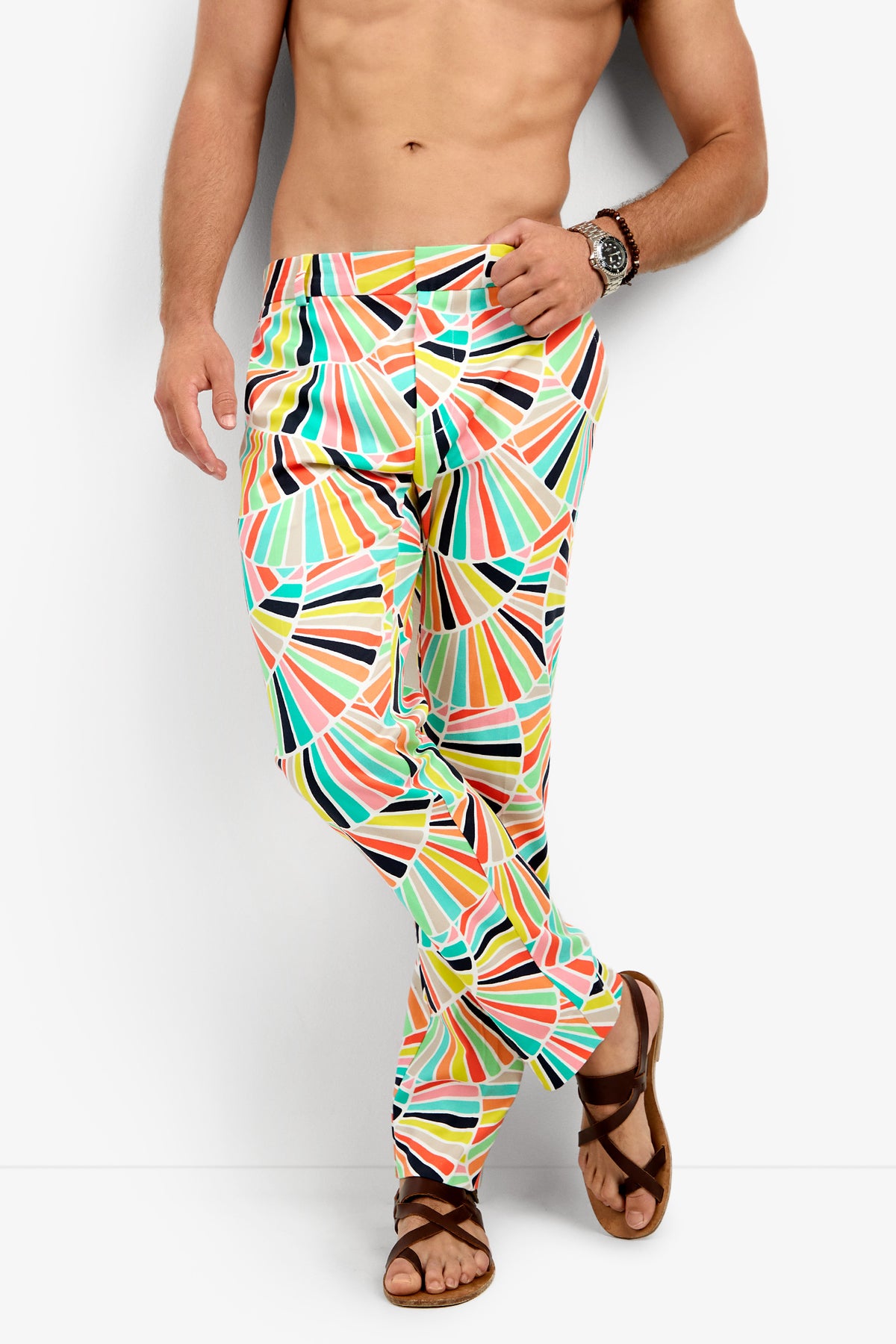A shirtless man wearing the Clyde Slim Trouser, featuring colorful geometric patterns, and brown sandals stands against a white background with one leg crossed over the other. He sports a wristwatch on his left wrist.