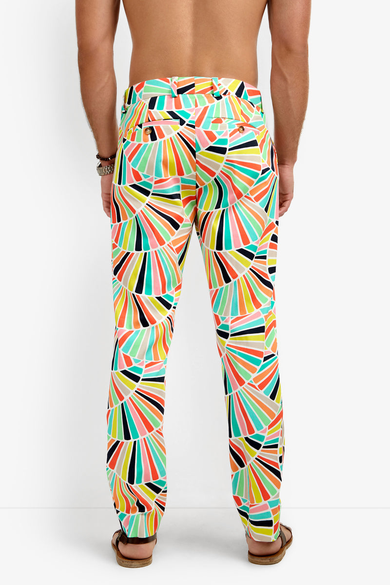 A shirtless person seen from the back wears Clyde Slim Trouser featuring a kaleidoscope of geometric shapes in orange, green, yellow, blue, pink, and black, paired with brown sandals.
