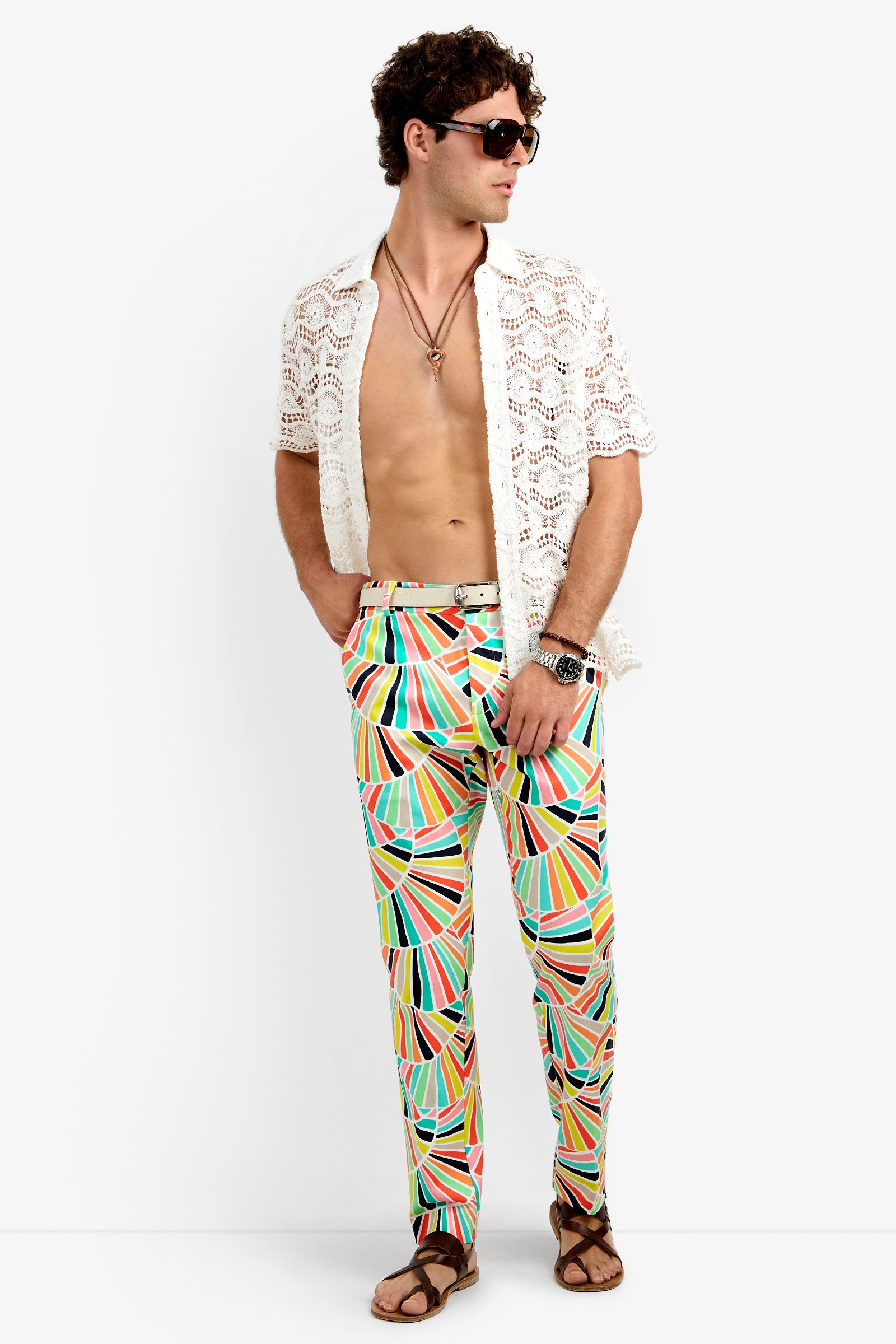 A man stands confidently with hands in his pockets against a plain background, wearing a white crochet short-sleeve shirt, Clyde Slim Trouser in a colorful geometric pattern, brown sandals, sunglasses, and necklaces.
