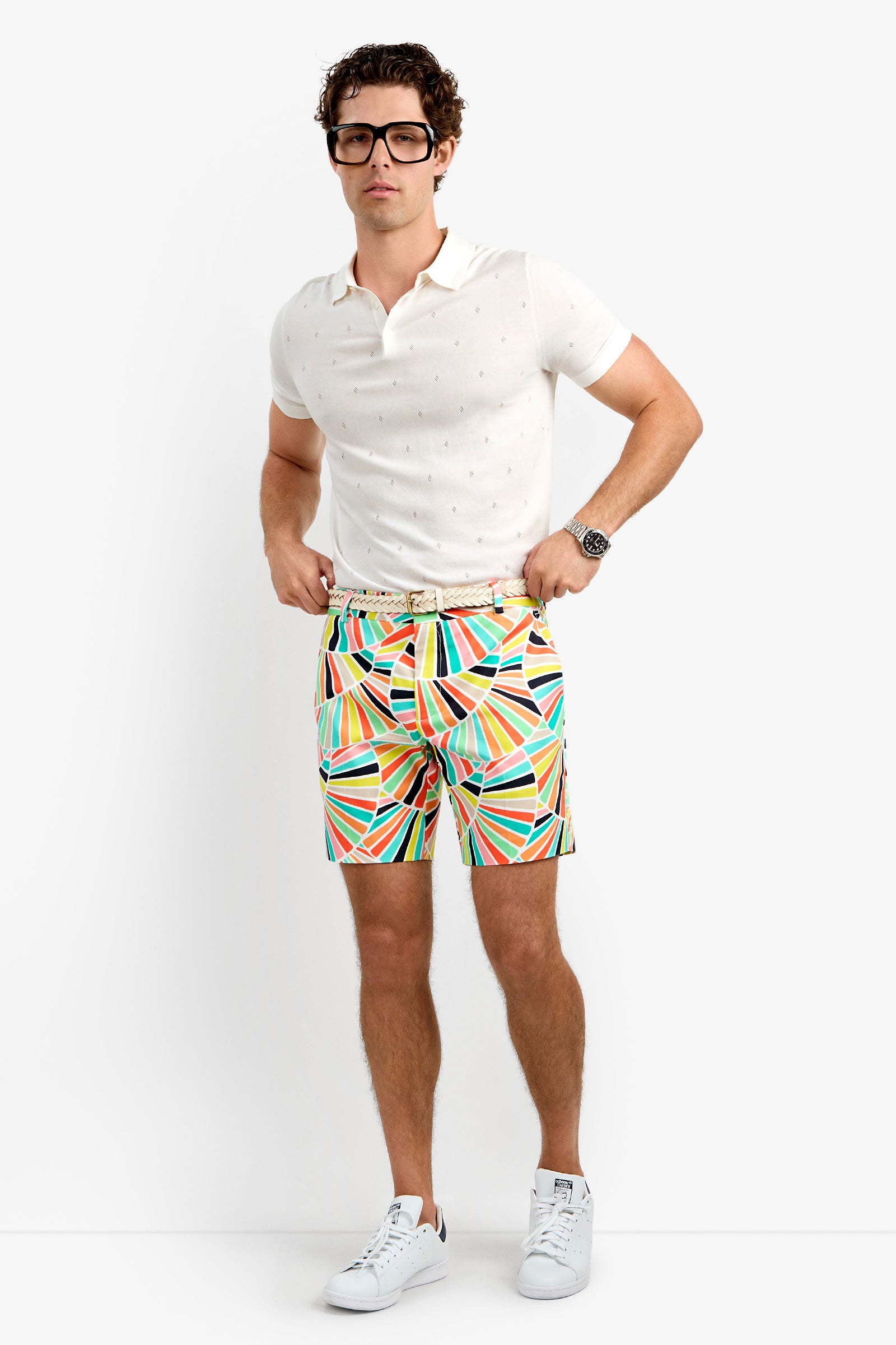A man stands against a plain background wearing large glasses, a white short-sleeve polo shirt, the colorful geometric Lawrence Short, a white belt, wristwatch, and white sneakers.