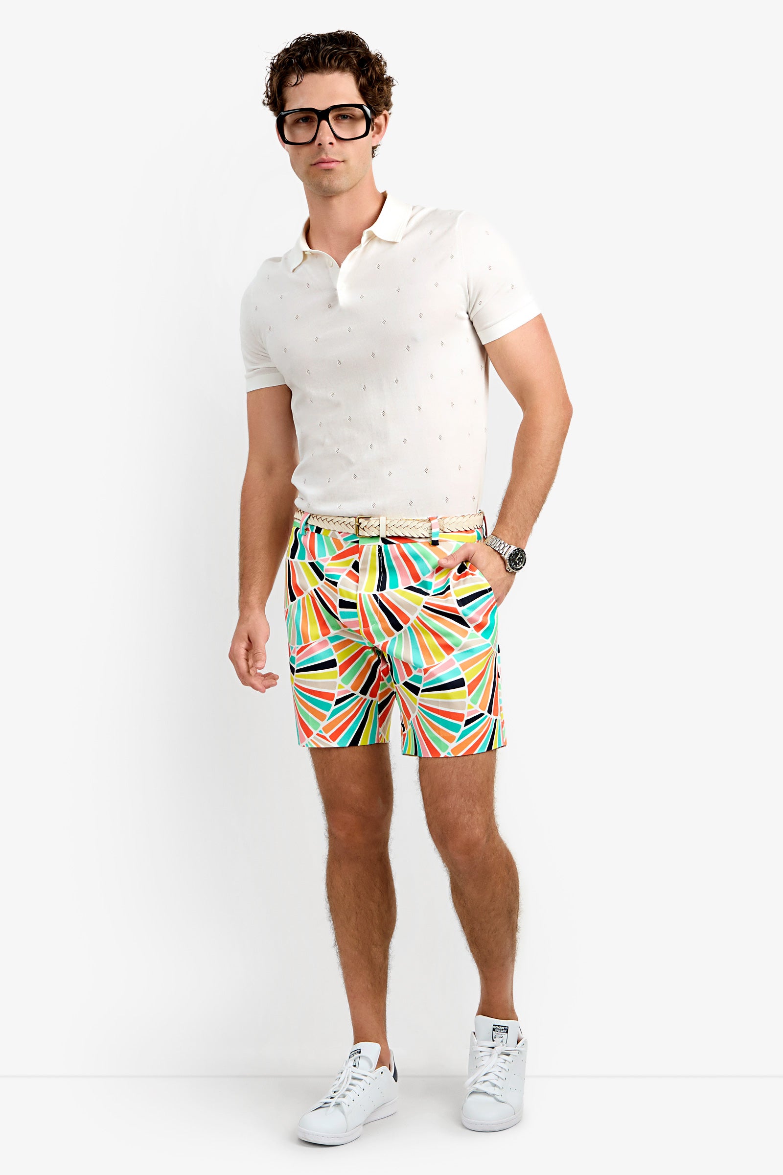 A man wearing a white polo, Lawrence Short with a colorful geometric pattern, white sneakers, black glasses, and a watch stands against a plain white background.