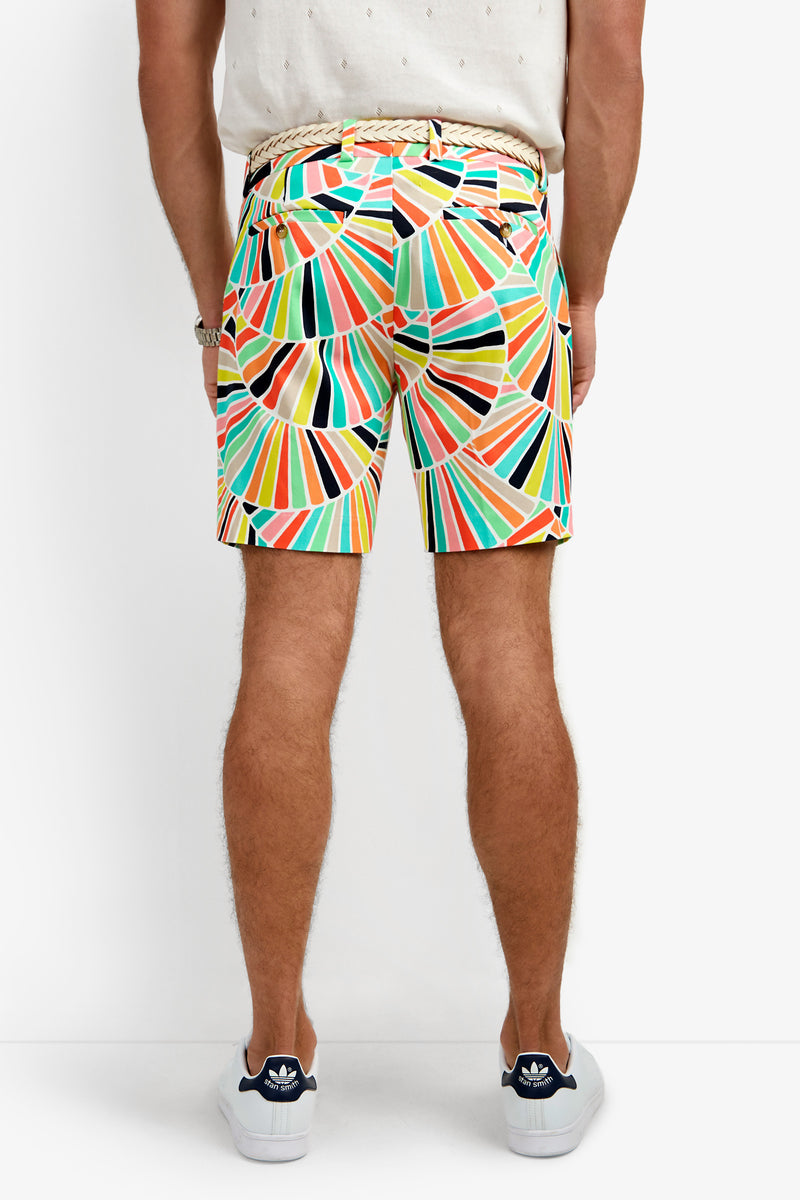 A person faces away, wearing the Lawrence Short with a colorful geometric pattern, a white shirt, beige woven belt, and white sneakers with navy accents against a plain white background.