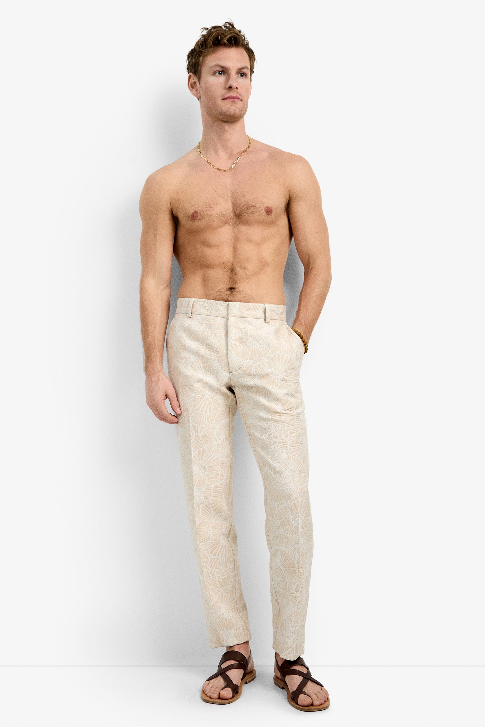A shirtless man with short hair and a necklace poses casually against a white background, wearing the Clyde Slim Trouser in Beachy Beige paired with brown sandals.