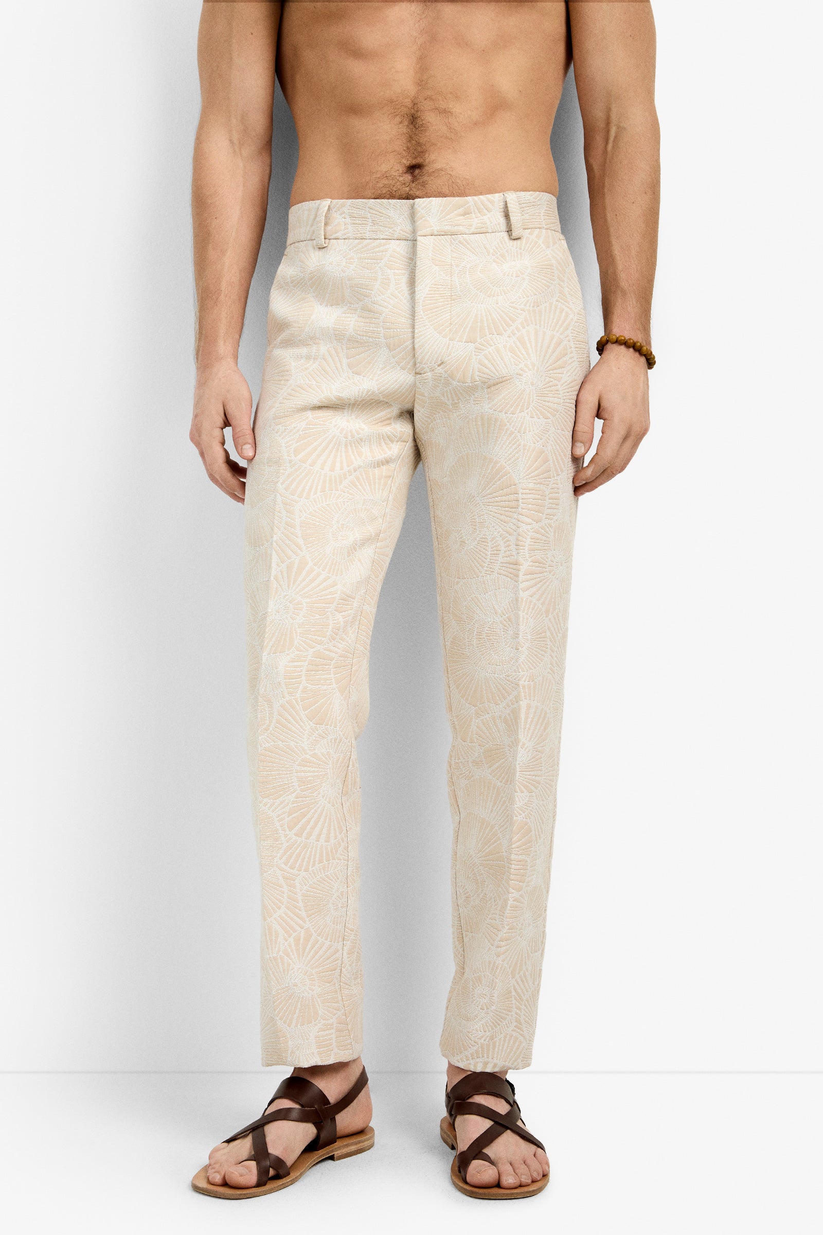 A shirtless man wearing Clyde Slim Trouser with a subtle leaf pattern, brown sandals, and a beaded bracelet stands against a plain white background.