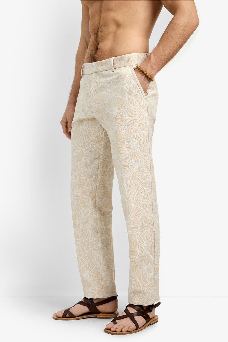 A shirtless man models the Clyde Slim Trouser in light beige with a subtle floral pattern, hands in pockets and wearing brown strappy sandals, standing against a white background.