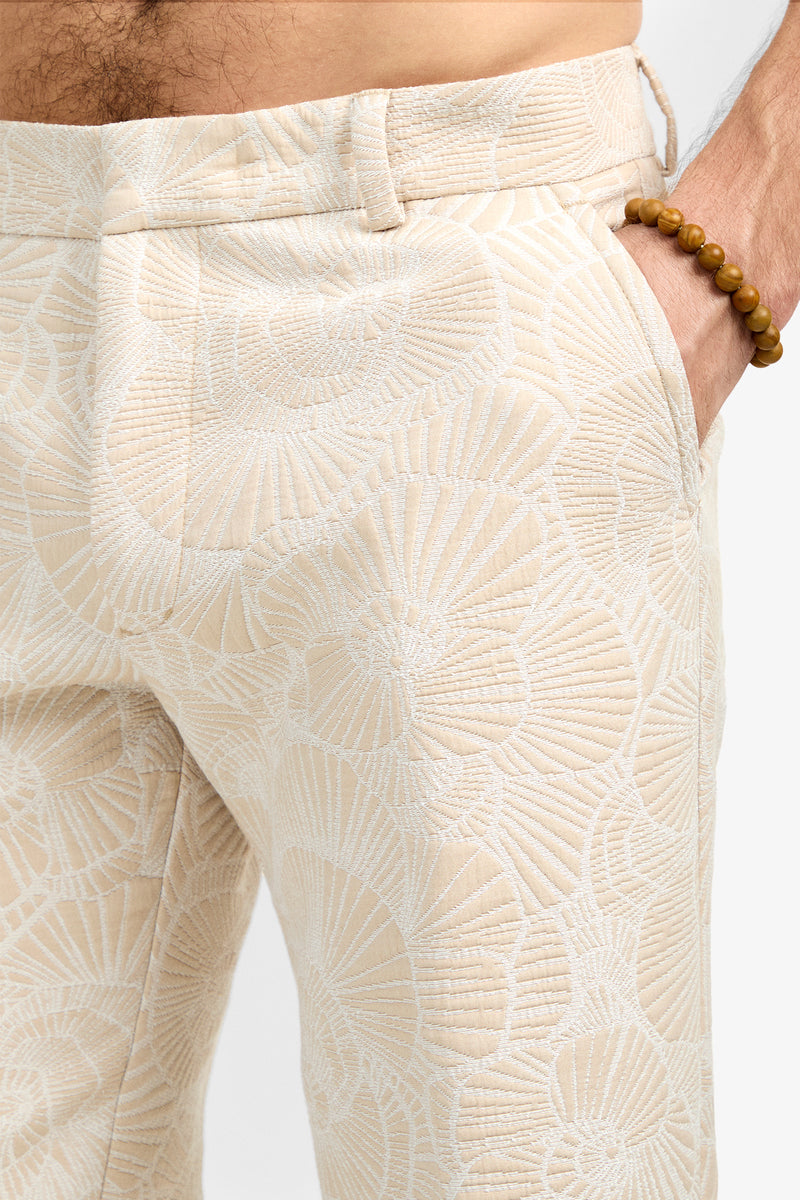 A close-up of a person wearing the Clyde Slim Trouser in cream with a textured, shell-like pattern. Their hand rests in the pocket, and they sport a beaded bracelet on their wrist.