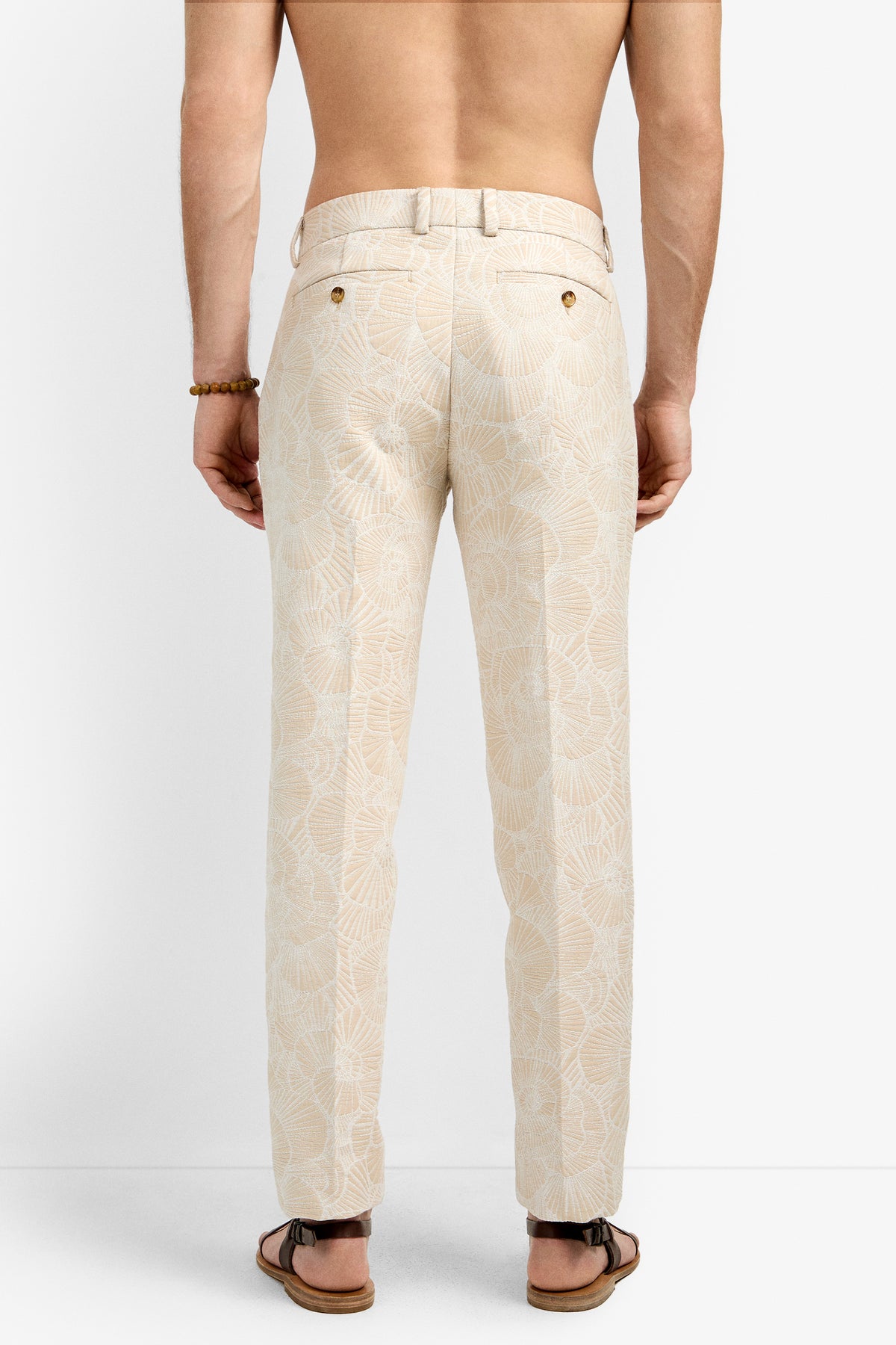 A shirtless person models the Clyde Slim Trouser, featuring beige fabric with a white leaf pattern, paired with a right wrist bracelet and brown sandals, photographed from the back against a plain background.