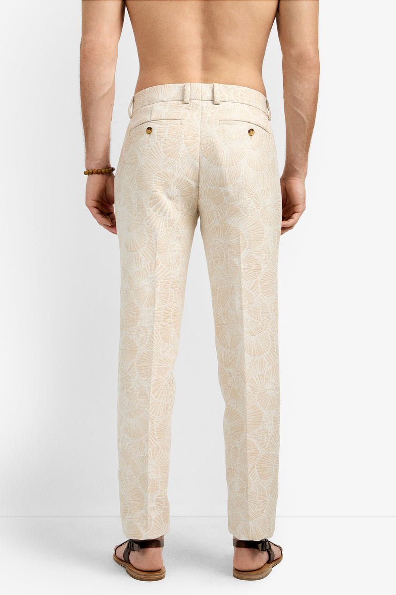 A shirtless person models the Clyde Slim Trouser, featuring beige fabric with a white leaf pattern, paired with a right wrist bracelet and brown sandals, photographed from the back against a plain background.