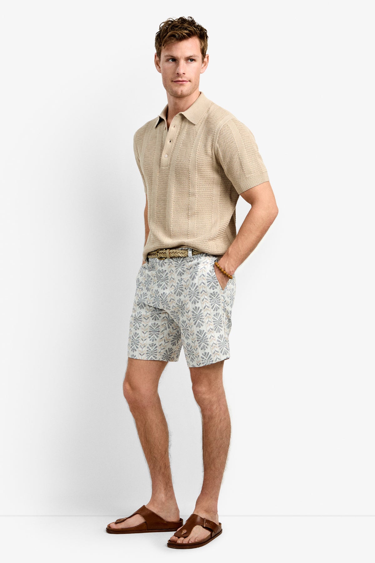 A man stands against a white background wearing beige short-sleeve knit polo, the Lawrence Short in patterned light blue and white, a belt, and brown sandals, with his hands in his pockets.