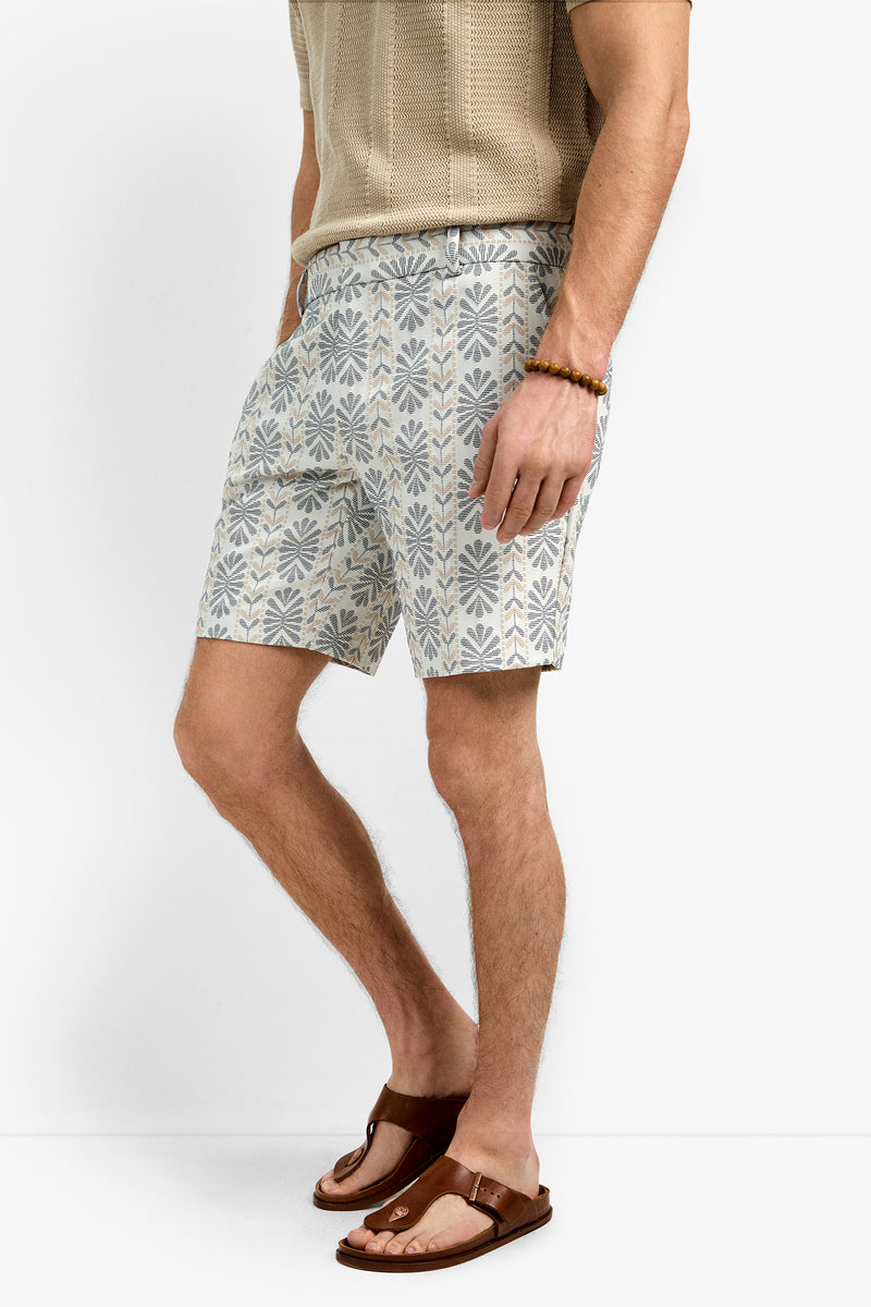 A man wearing the Lawrence Short, a beige short-sleeve knit shirt, brown sandals, and a beaded bracelet stands against a plain white background.