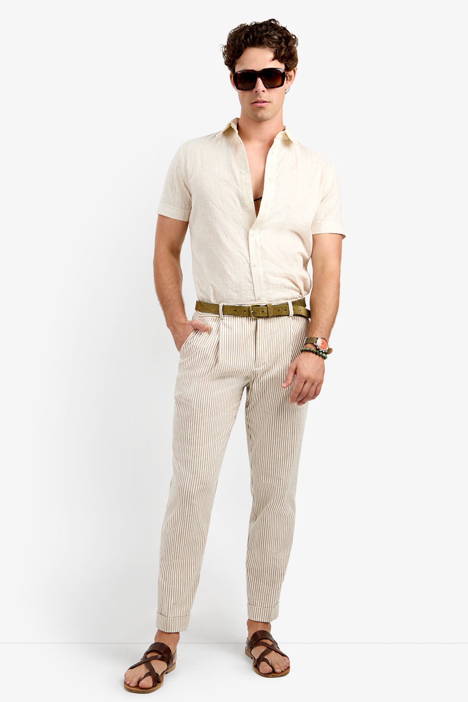 A man stands against a white background wearing the Beale Pleated Trouser, a light short-sleeve button-up shirt, brown belt, brown sandals, sunglasses, and a beaded bracelet, with his hands in his pockets.