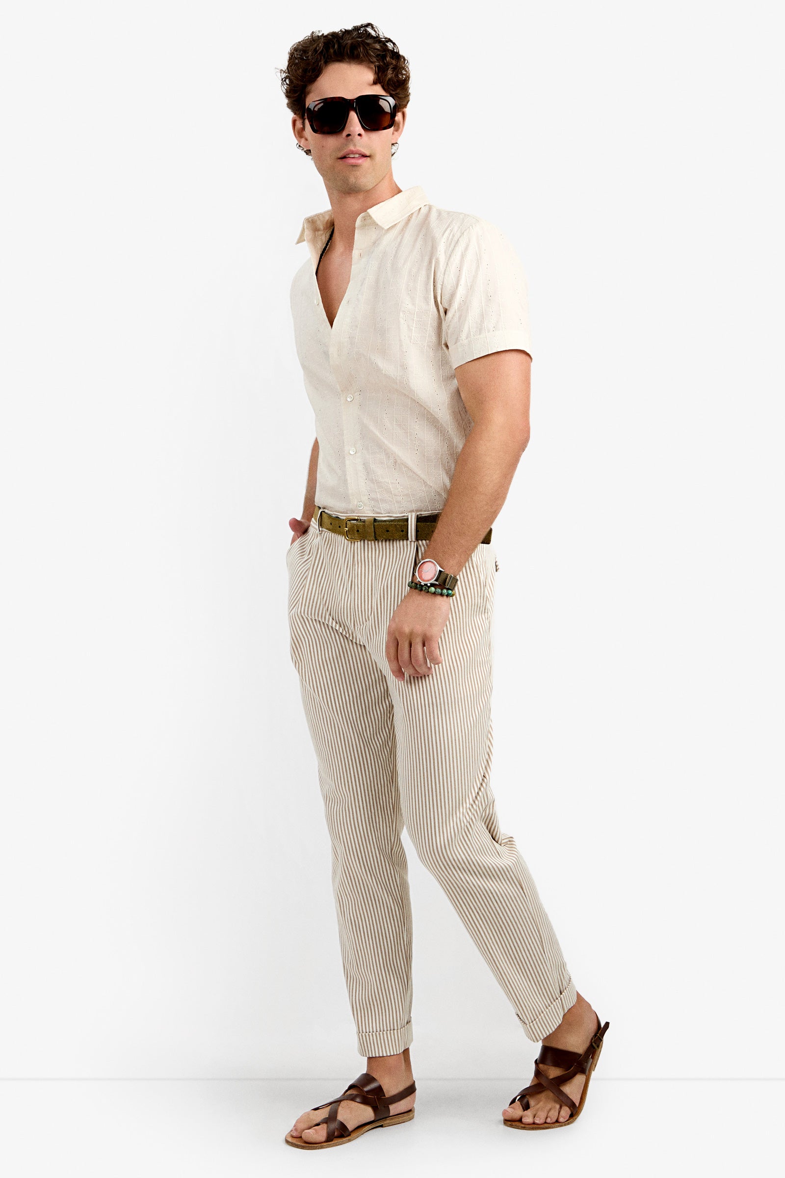 A man wearing sunglasses, a short-sleeve beige button-up shirt, Beale Pleated Trouser in striped beige, a brown belt, a watch, and brown sandals stands against a plain white background.