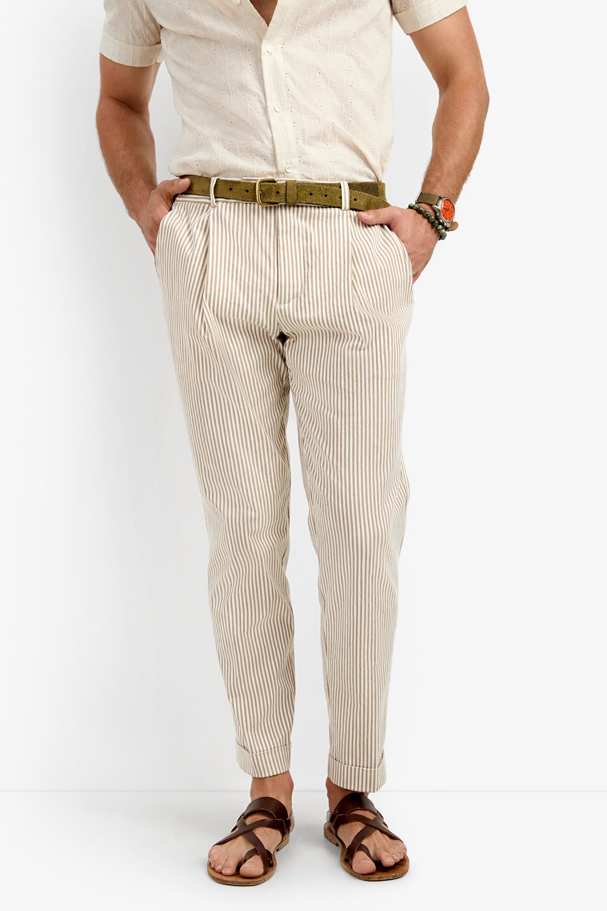 A person, shown from shoulders down, wears the Beale Pleated Trouser—a tan and white striped pant—paired with a light short-sleeve shirt, brown belt, brown sandals, and beaded bracelets. Hands are in pockets.