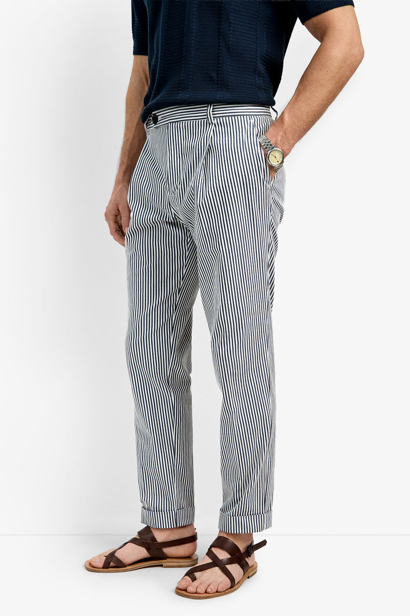 A person wearing navy short sleeves, Beale Pleated Trousers in blue-and-white vertical stripes, brown sandals, and a wristwatch, standing against a white background.