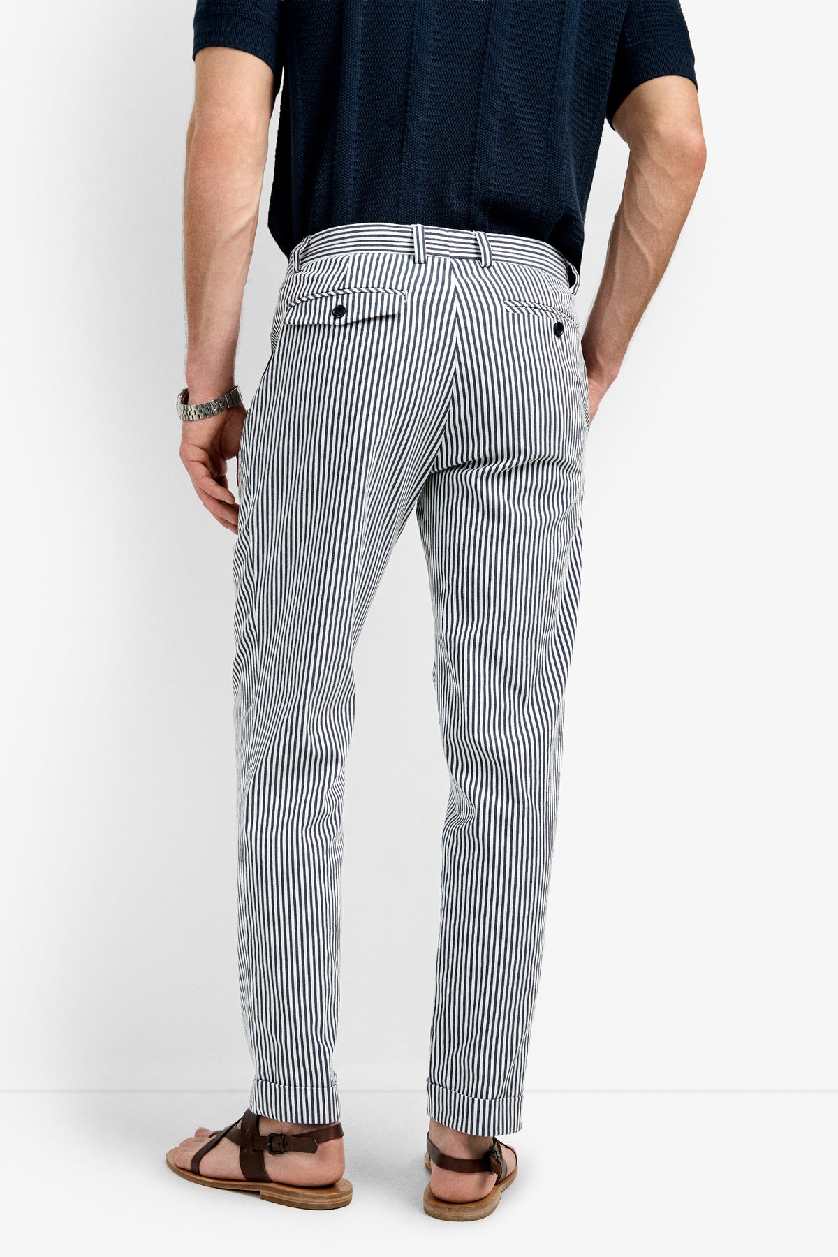 Wearing a navy short-sleeve shirt, Beale Pleated Trouser in white and navy vertical stripes, brown watch, and brown sandals, a person stands with their back to the camera against a plain white background.