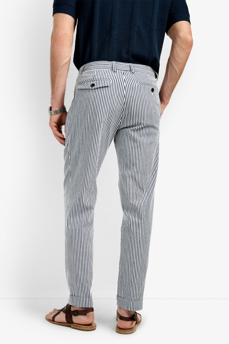 Wearing a navy short-sleeve shirt, Beale Pleated Trouser in white and navy vertical stripes, brown watch, and brown sandals, a person stands with their back to the camera against a plain white background.