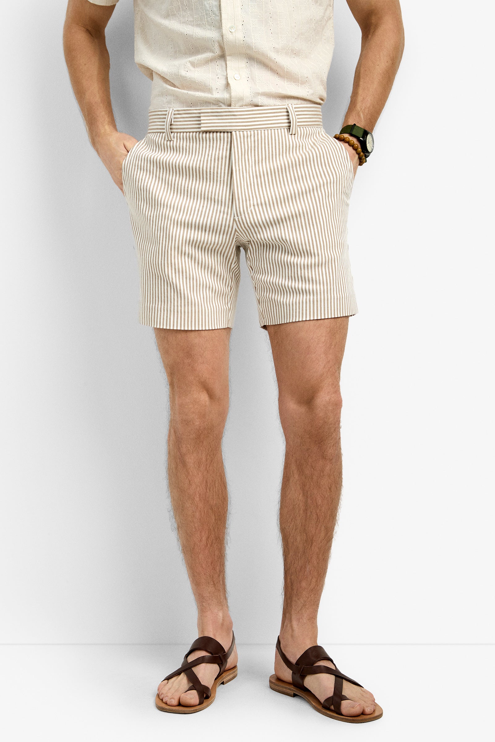 A man wearing the Roger Short, beige and white vertically striped shorts, pairs them with a light short-sleeve shirt, brown sandals, and a wristwatch while standing against a plain white background with hands in his pockets.