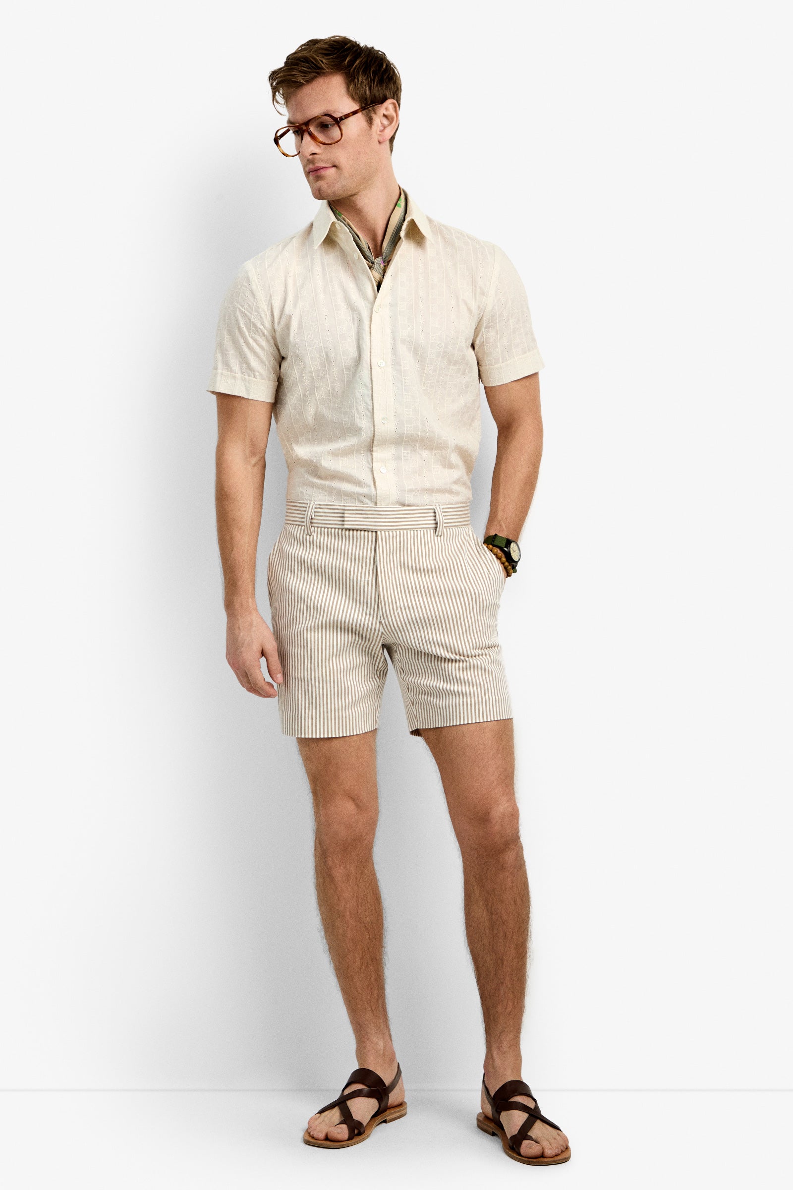 A man with a neutral expression stands against a white background wearing glasses, a beige short-sleeve shirt, Roger Short, brown sandals, a watch, and has his hands in his pockets.