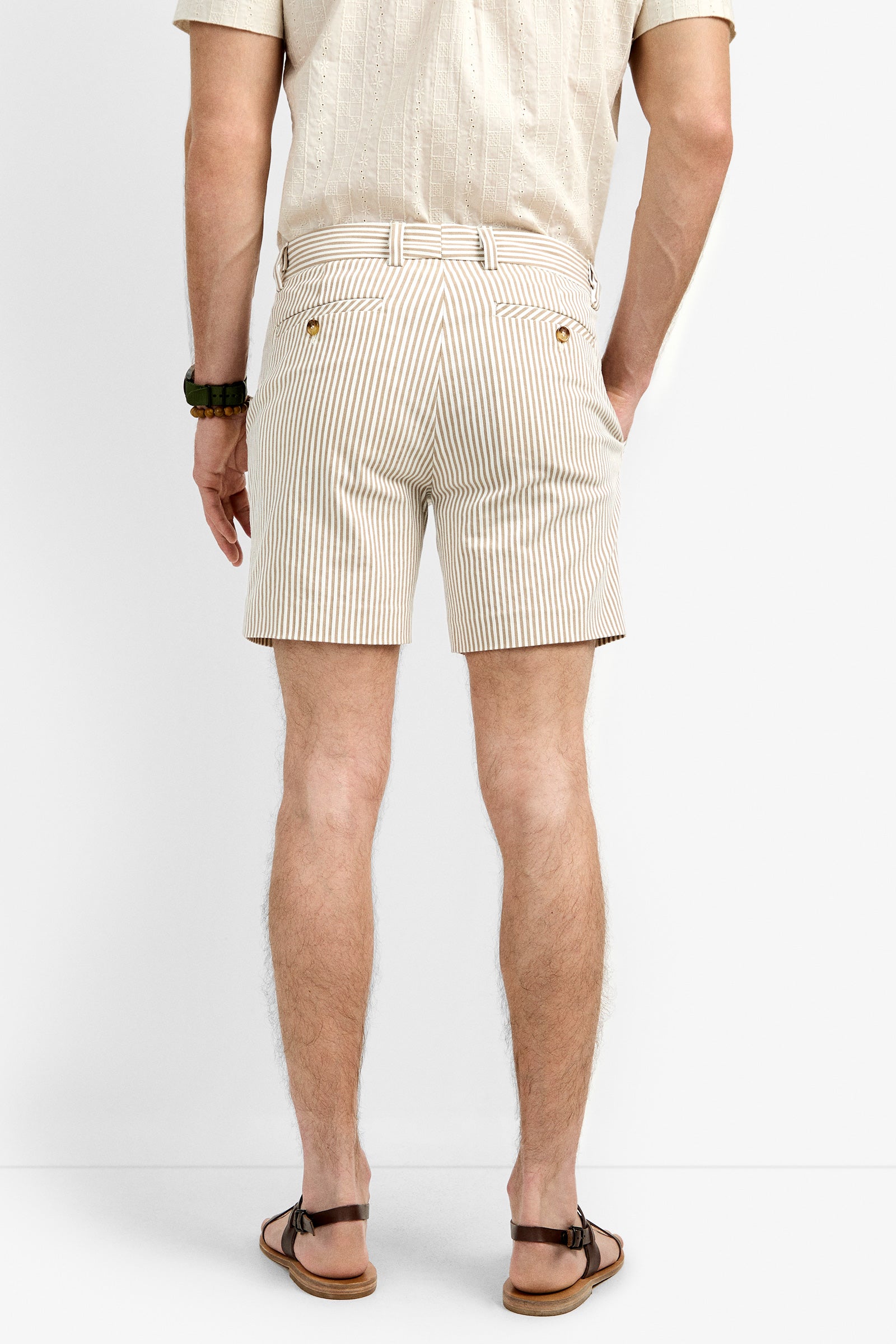 A man stands with his back to the camera in beige and white striped Roger Shorts, a light short-sleeve shirt, brown sandals, and a beaded bracelet against a plain white backdrop.