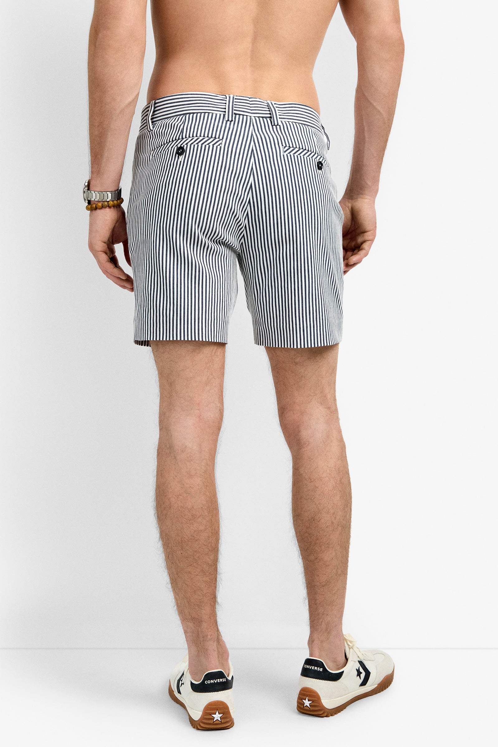 A shirtless man faces away from the camera, wearing Roger Short featuring vertical stripes and white sneakers with black accents. His arms hang relaxed by his sides, and a watch is visible on his left wrist.